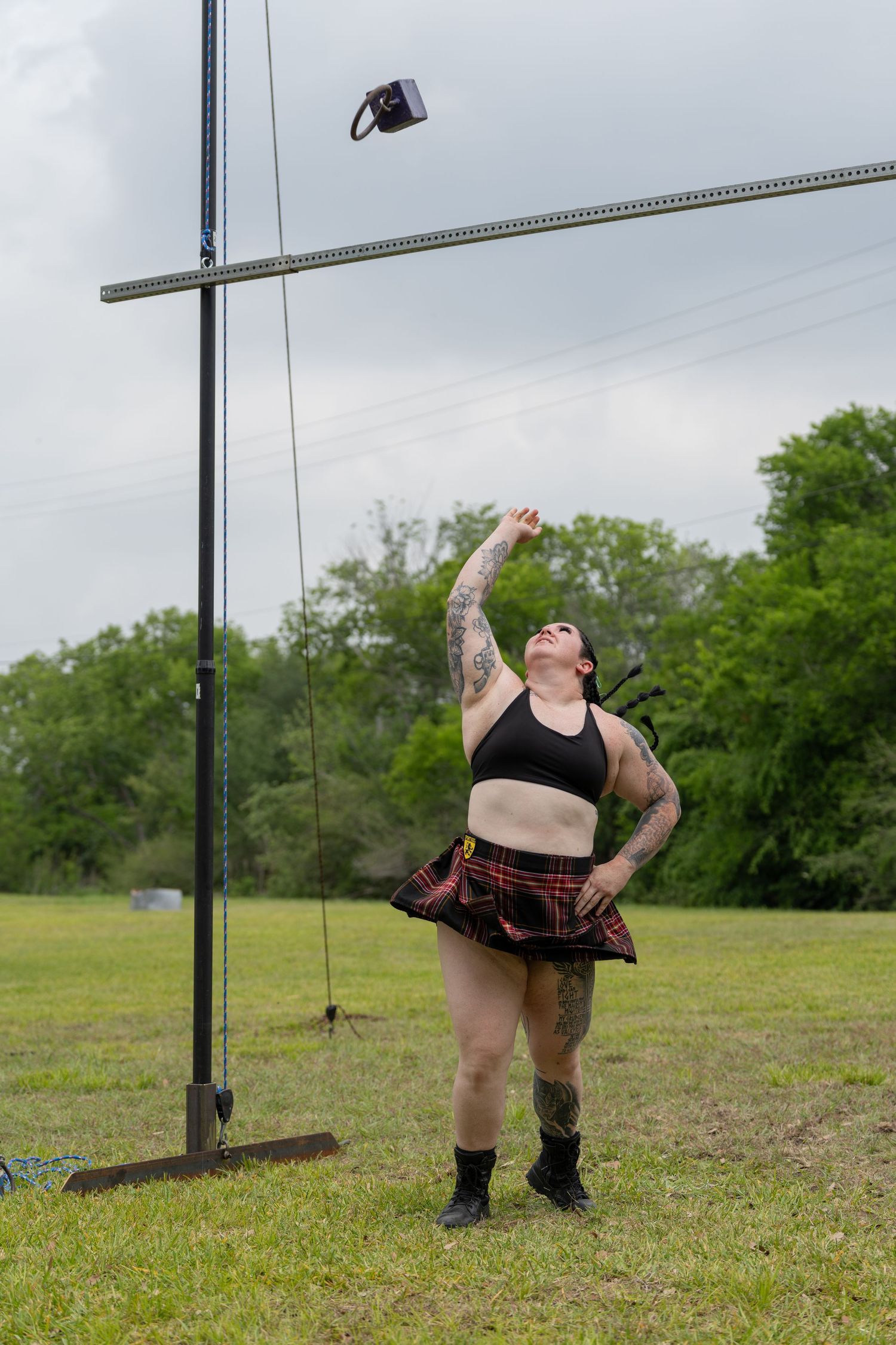 Tattooed woman in plaid skirt and crop top tosses object skyward at outdoor Highland Games competition.