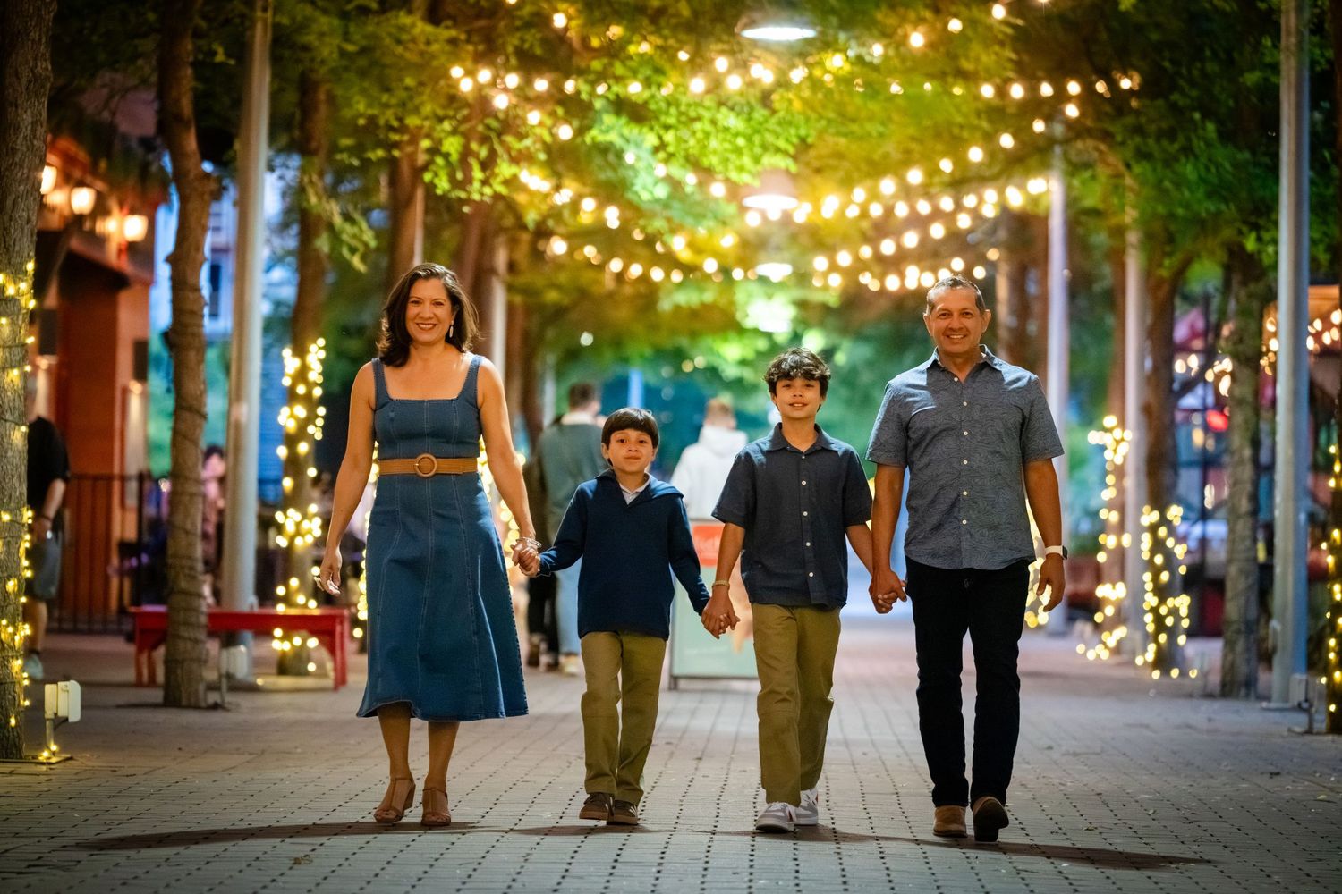 Family of four walking hand-in-hand on a festive lit pathway at night with twinkling string lights overhead.