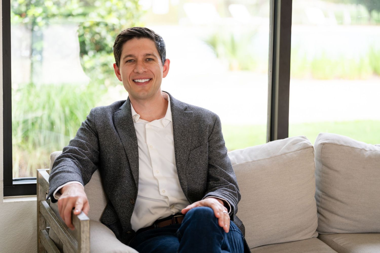 Smiling man in gray blazer sitting relaxed on a white sofa in a bright, modern living room.
