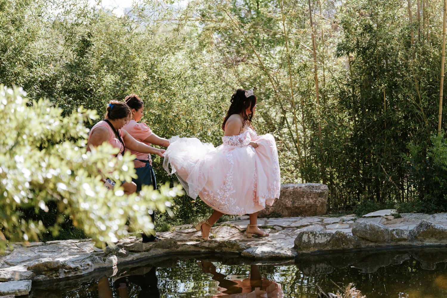 Bride in pink wedding gown steps across garden pond rocks assisted by attendants amid lush green trees.