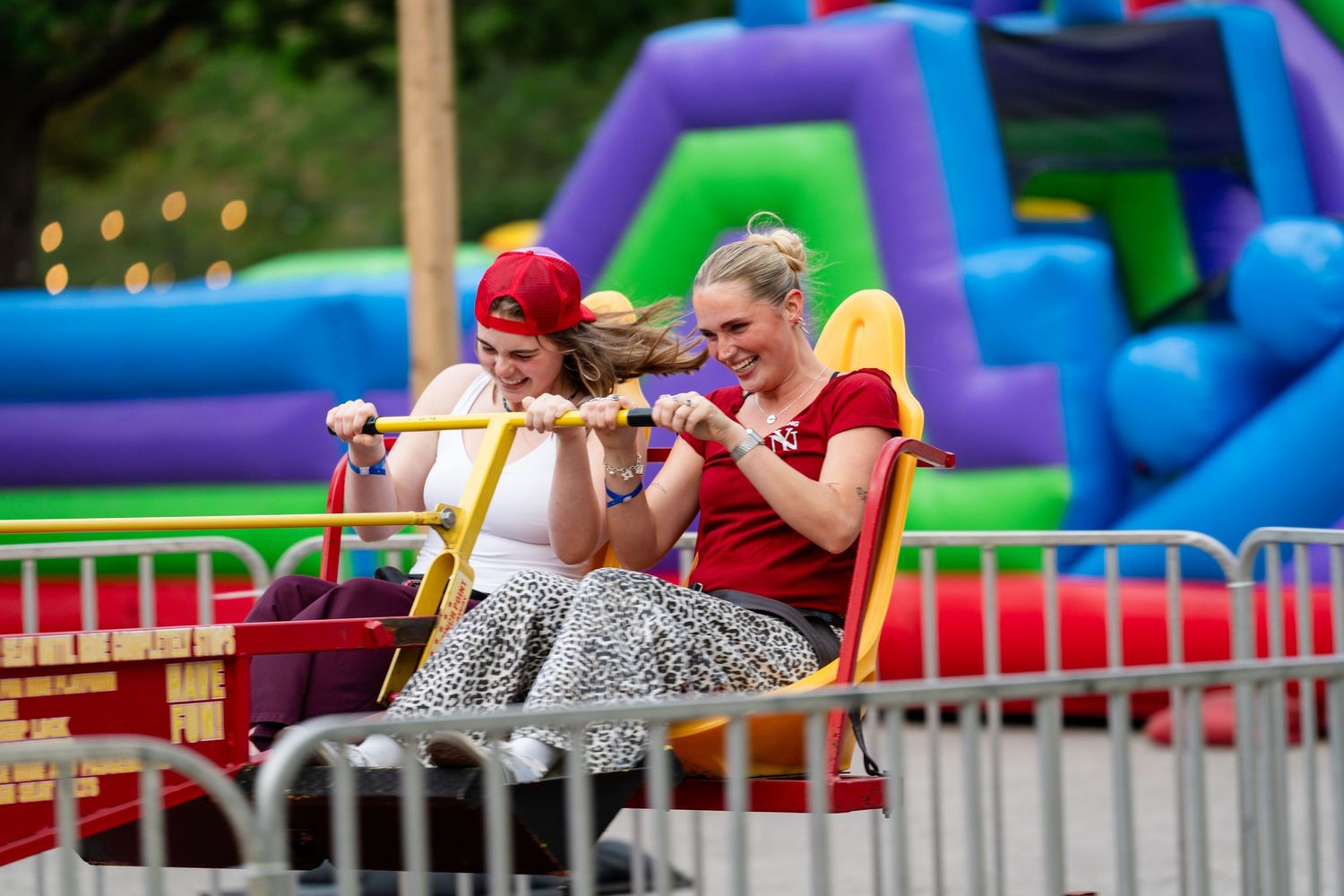 Two women laughing and holding on while riding a colorful carnival ride at an outdoor fair.