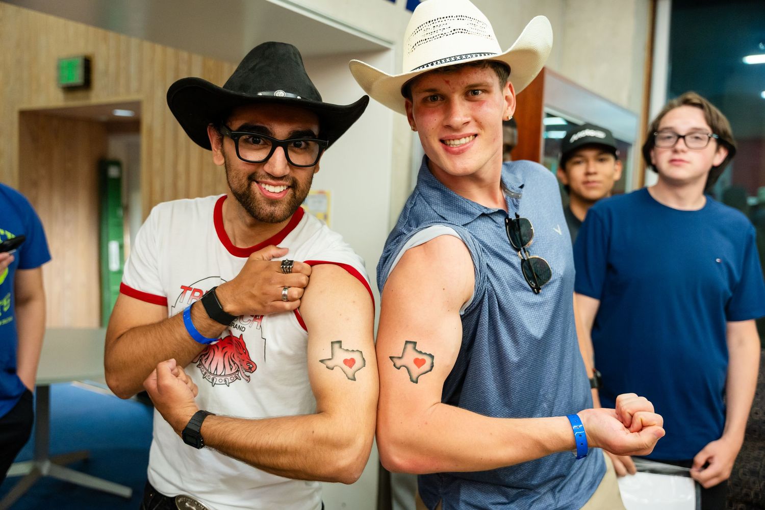 Two men in cowboy hats showing matching Texas-shaped tattoos on their arms at an indoor event.