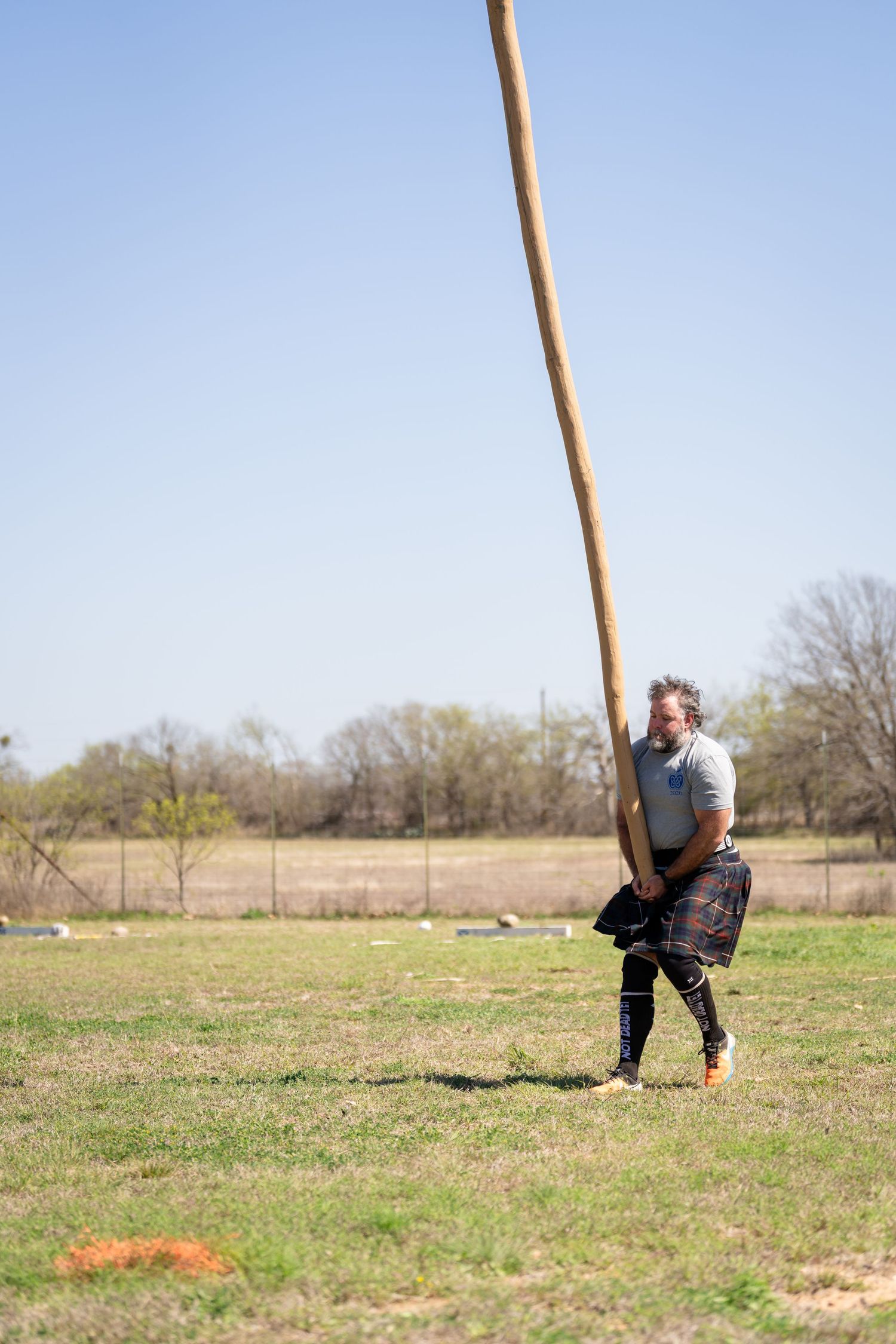 Man in kilt tossing a caber at a Scottish Highland Games competition on a sunny day.