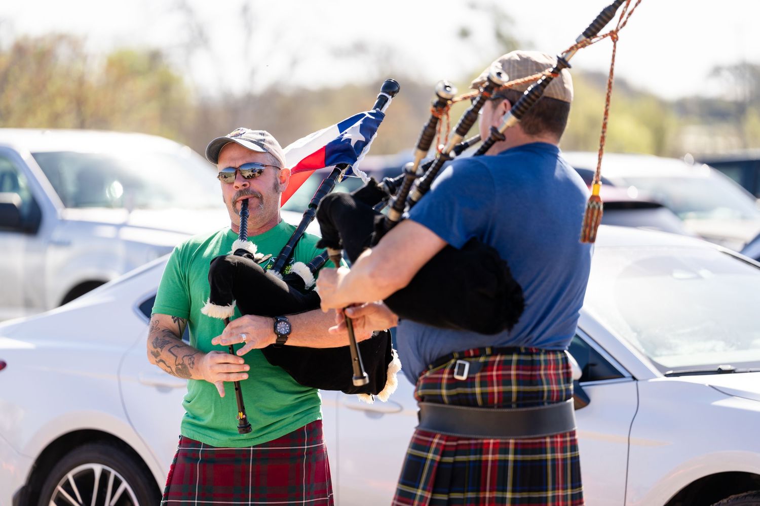 Two men in kilts playing bagpipes outdoors at a Scottish festival, with cars and a flag visible in the background.