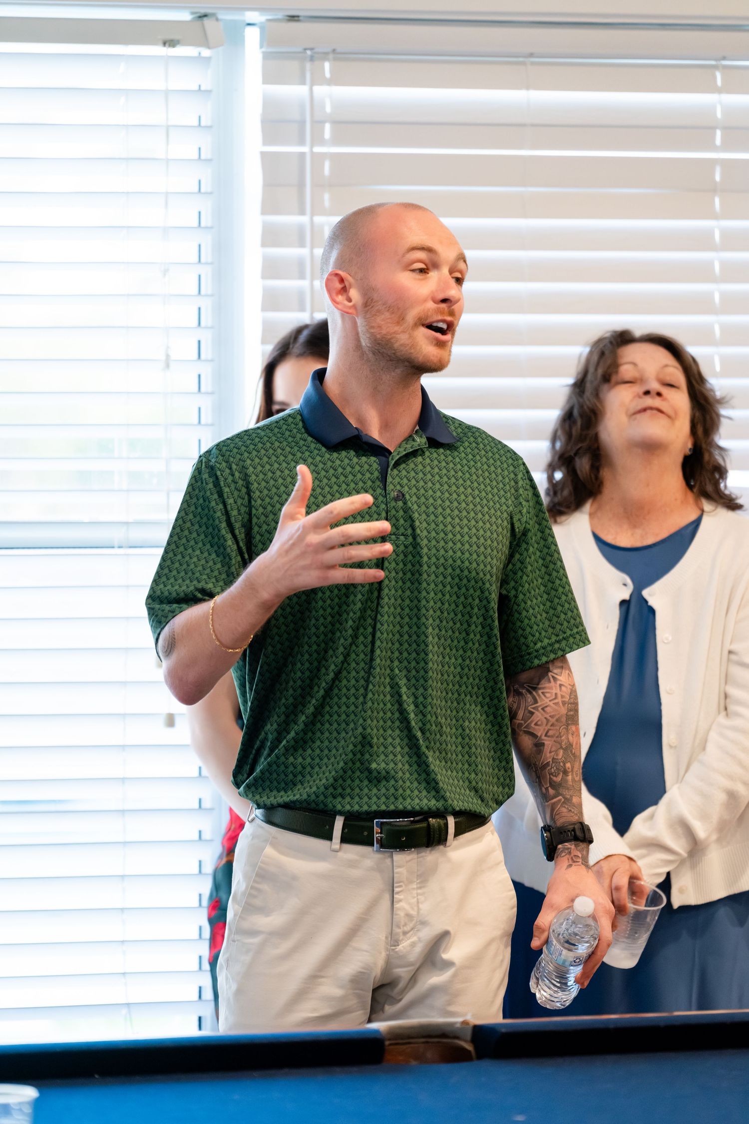 Tattooed bald man in green polo gestures while speaking, woman in white cardigan holds water bottle behind him.