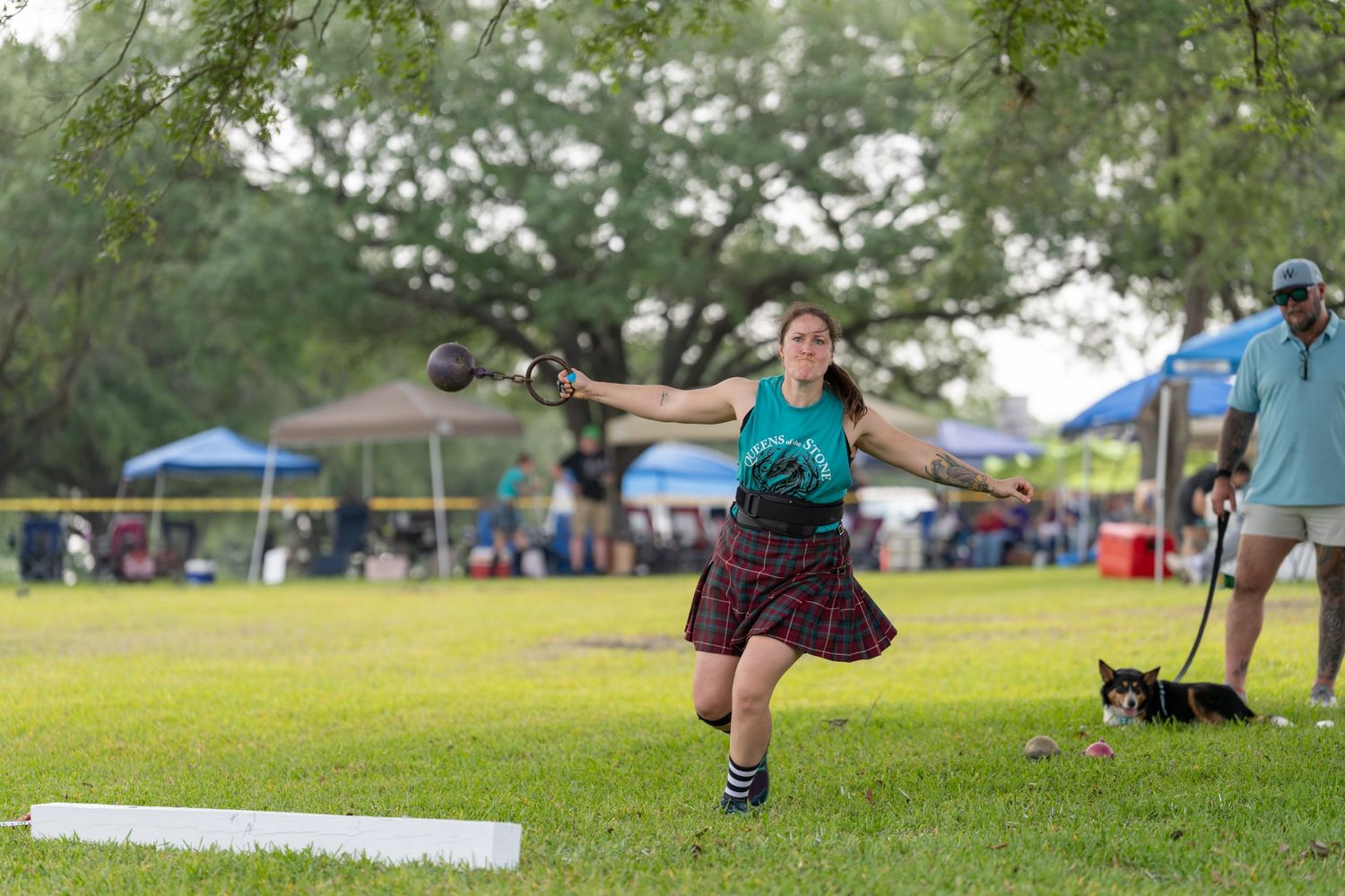 Woman in tartan skirt competes in Highland Games hammer throw on green field with spectators and tents in background.