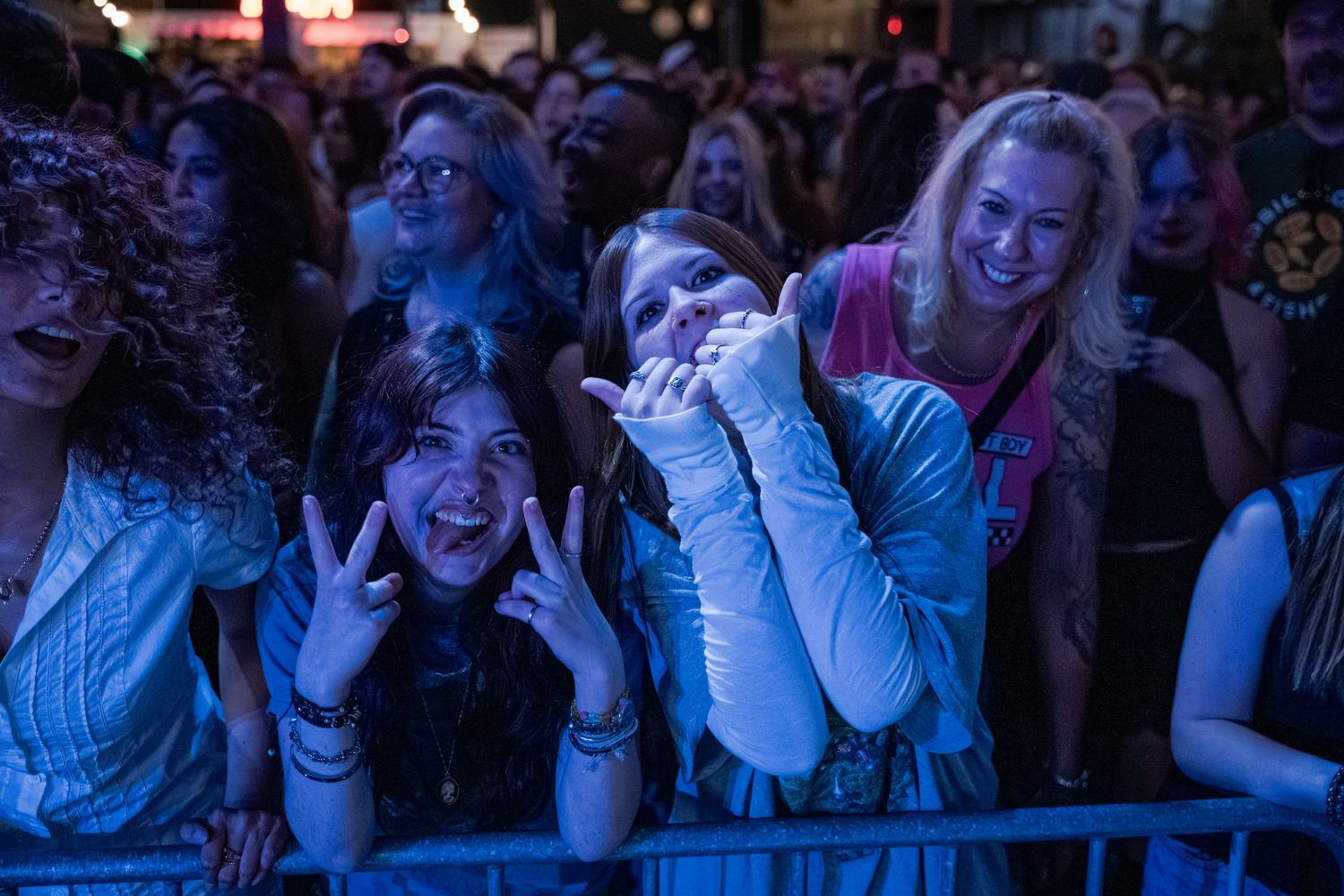Excited concert crowd cheering at outdoor music festival, fans smiling and making peace signs behind barrier.
