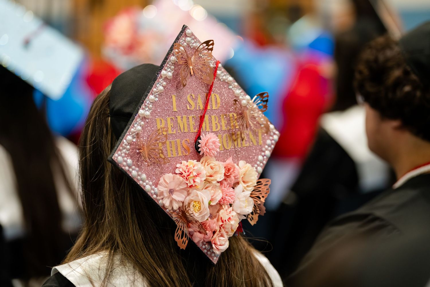 Decorated graduation cap with pink flowers and gold text at a commencement ceremony.