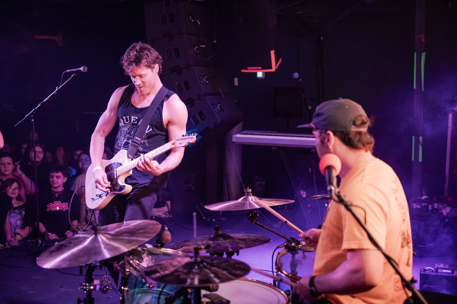 Two musicians perform live on stage, one playing electric guitar and another on drums, with a crowd visible in the background.