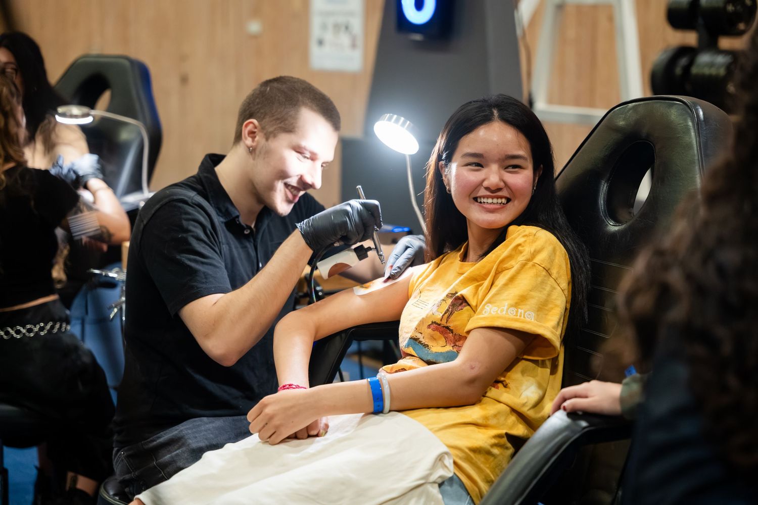 Tattoo artist tattooing a smiling young woman in a yellow shirt at a tattoo studio.
