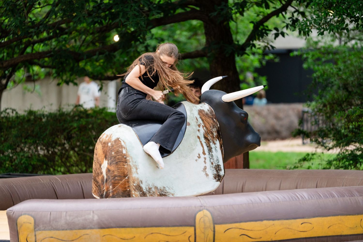 Woman riding a mechanical bull at an outdoor event, surrounded by inflatable padding and green trees in the background.
