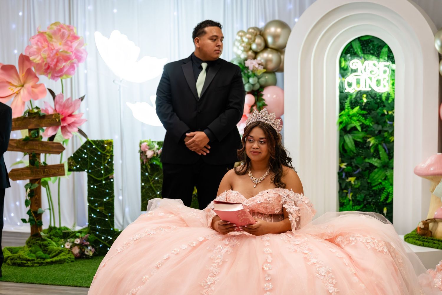 Young woman in pink quinceañera gown seated at decorated table with male escort standing behind her at elegant party.