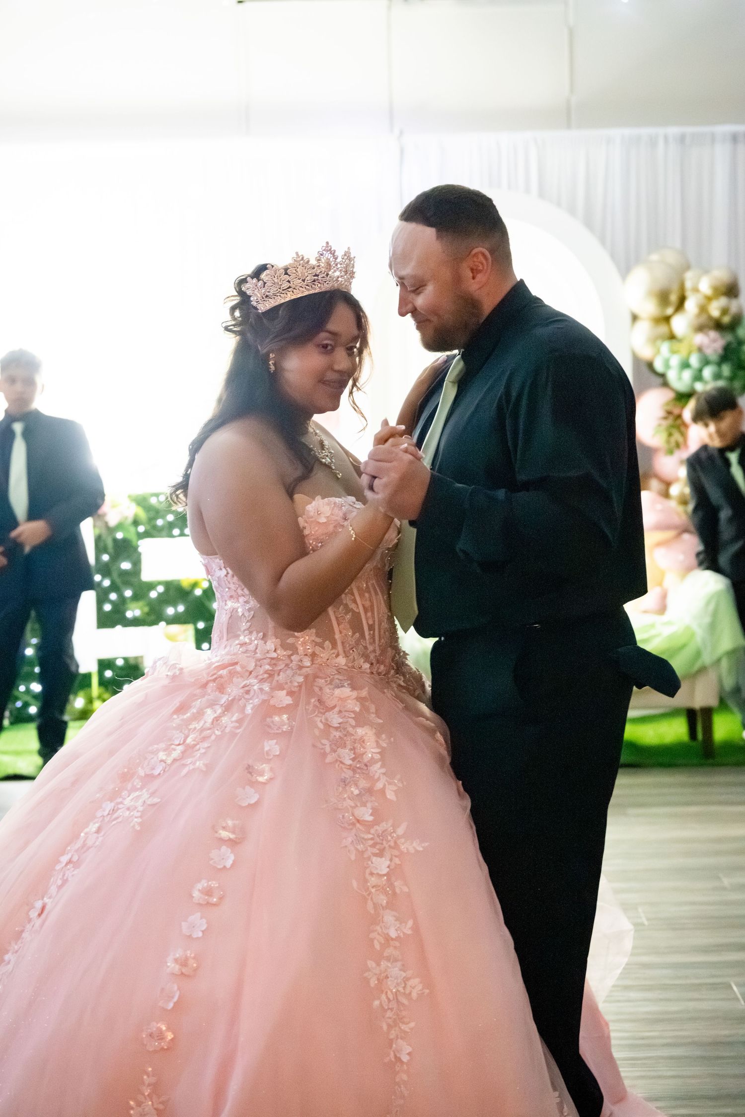 A young woman in a pink ball gown and tiara shares a dance with a man in a black suit at a quinceañera celebration.