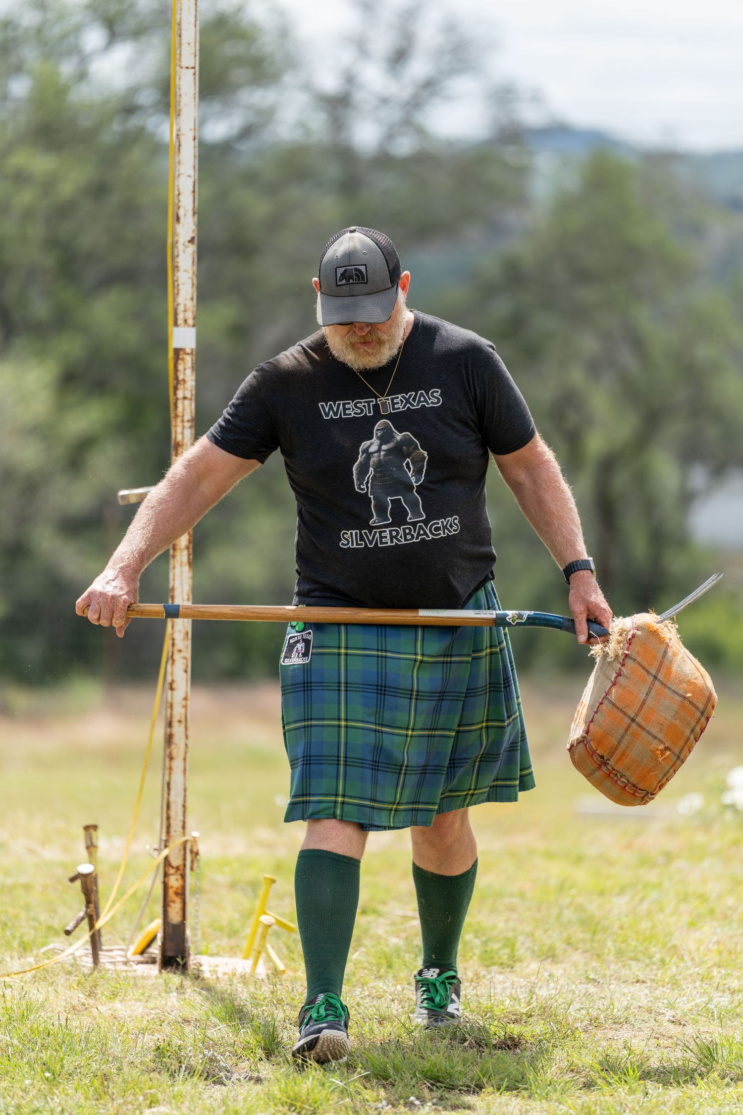 Man in green tartan kilt and West Texas Silverback shirt competing at Highland Games outdoor event.