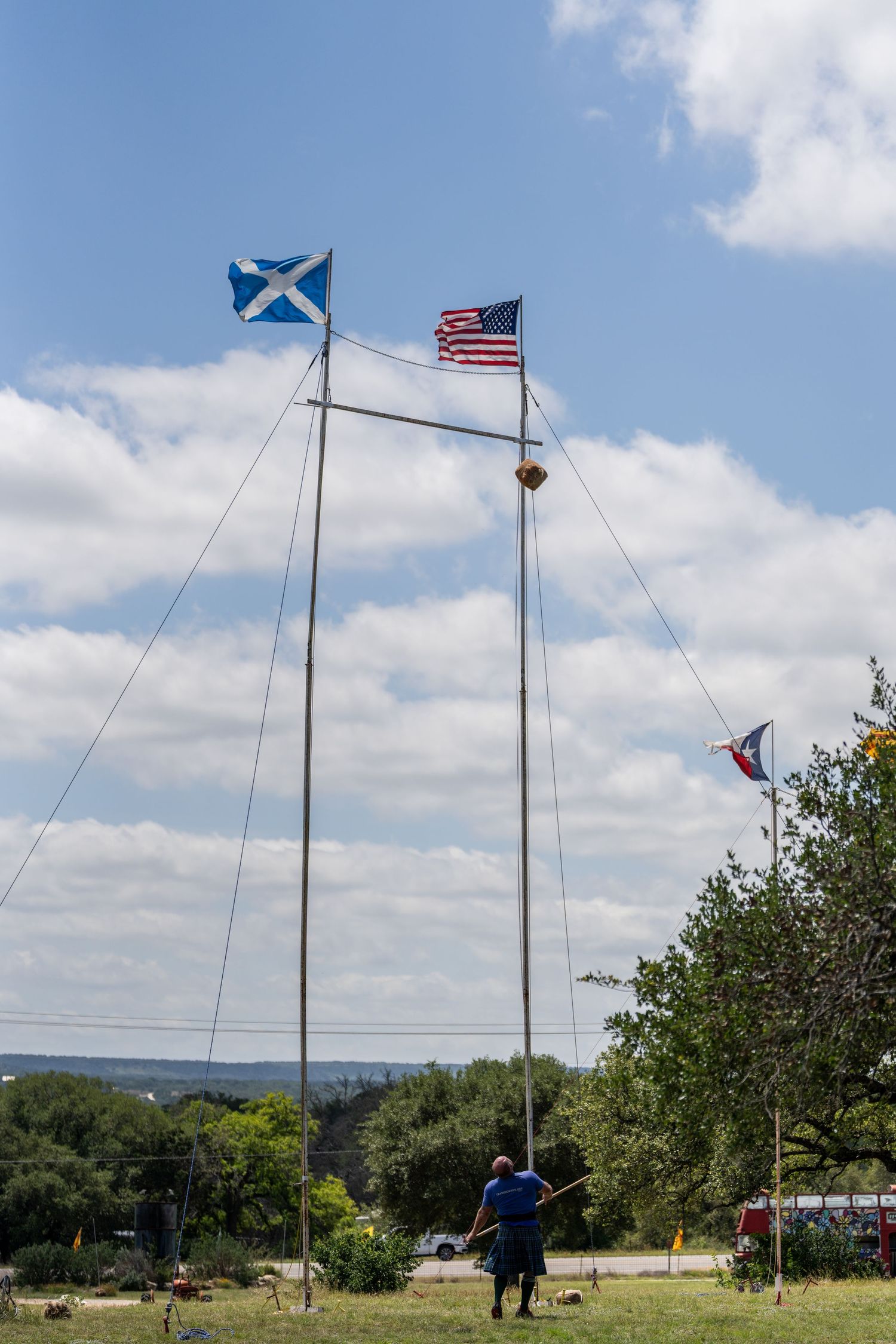 Three flagpoles flying Scottish, American, and Texas flags outdoors at a Highland Games festival on a sunny day.