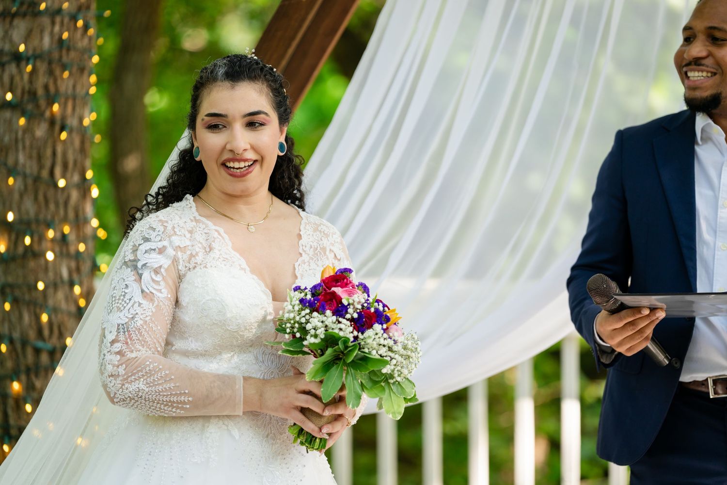 Smiling bride in lace wedding dress holding colorful bouquet during outdoor ceremony with white drape backdrop.