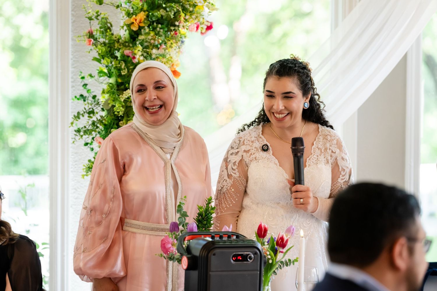 Two women smile joyfully at a wedding ceremony, one in a hijab and pink dress, the other in a white bridal gown holding a mic.
