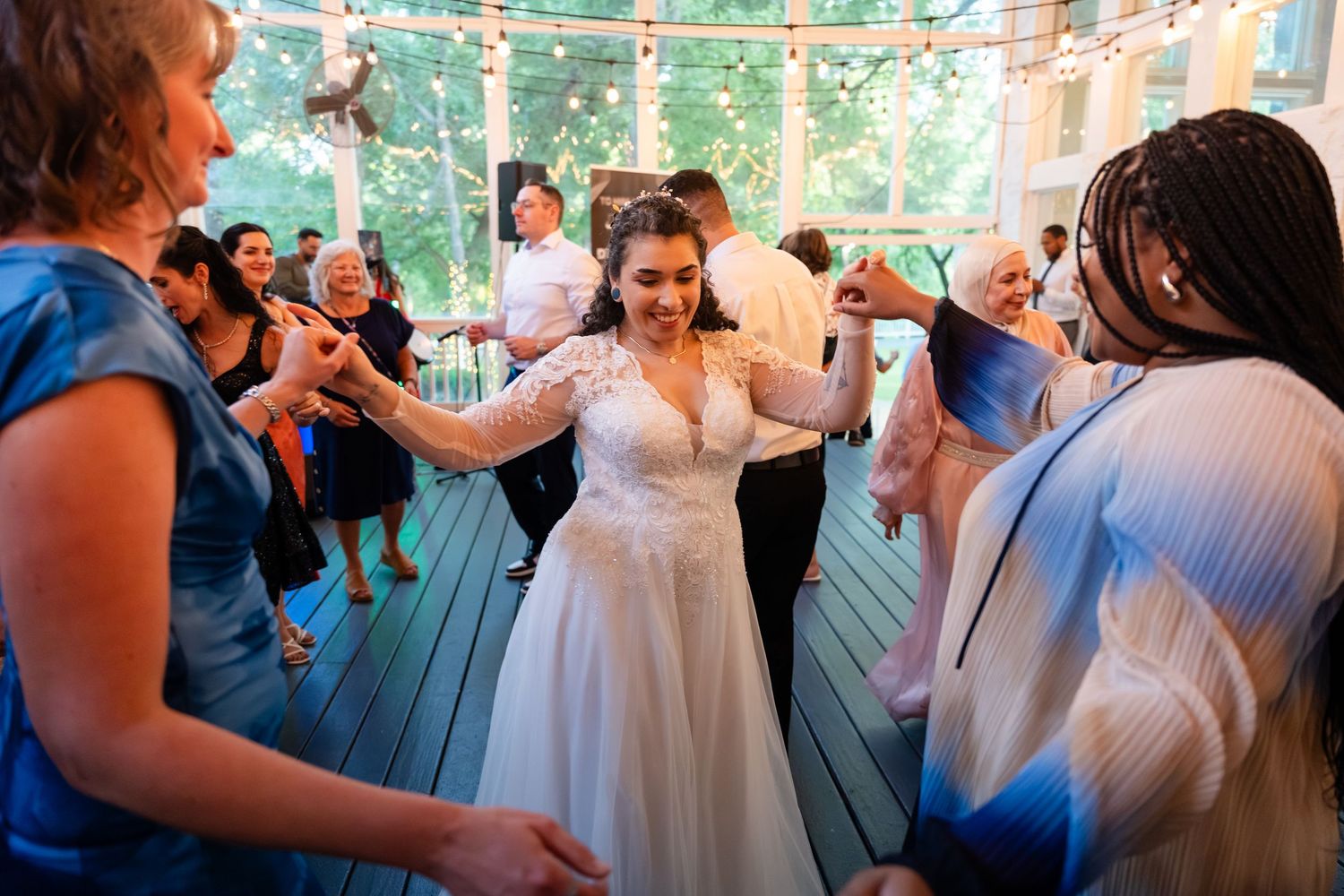 Bride in white lace gown dances joyfully with guests at indoor wedding reception with string lights.