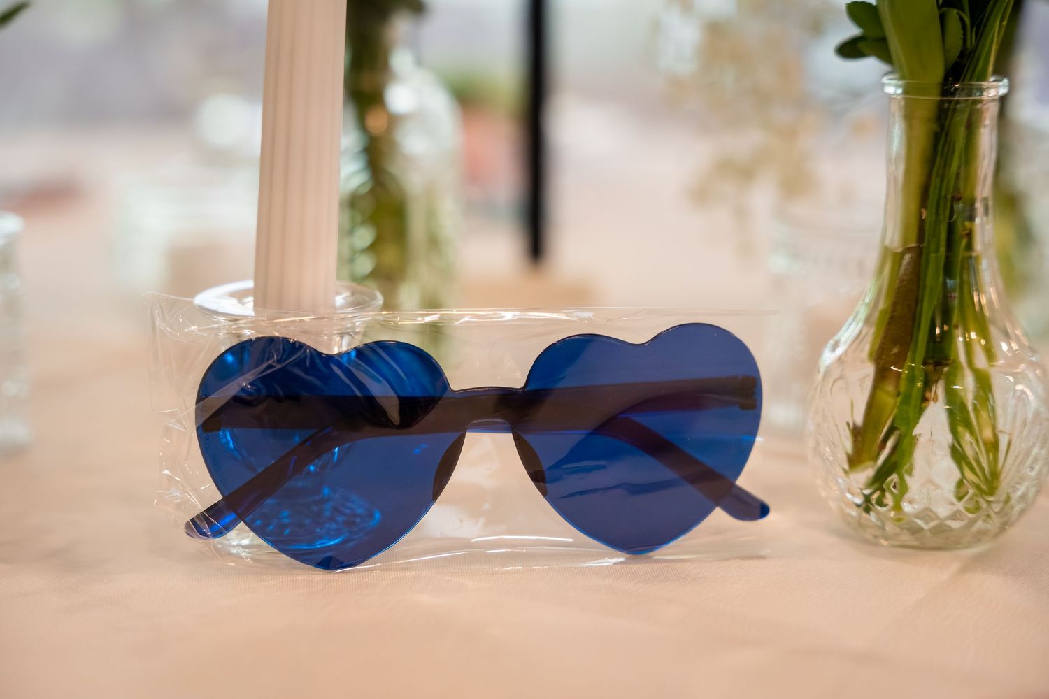 Blue heart-shaped sunglasses in clear packaging displayed on a table with floral arrangements at a wedding reception.