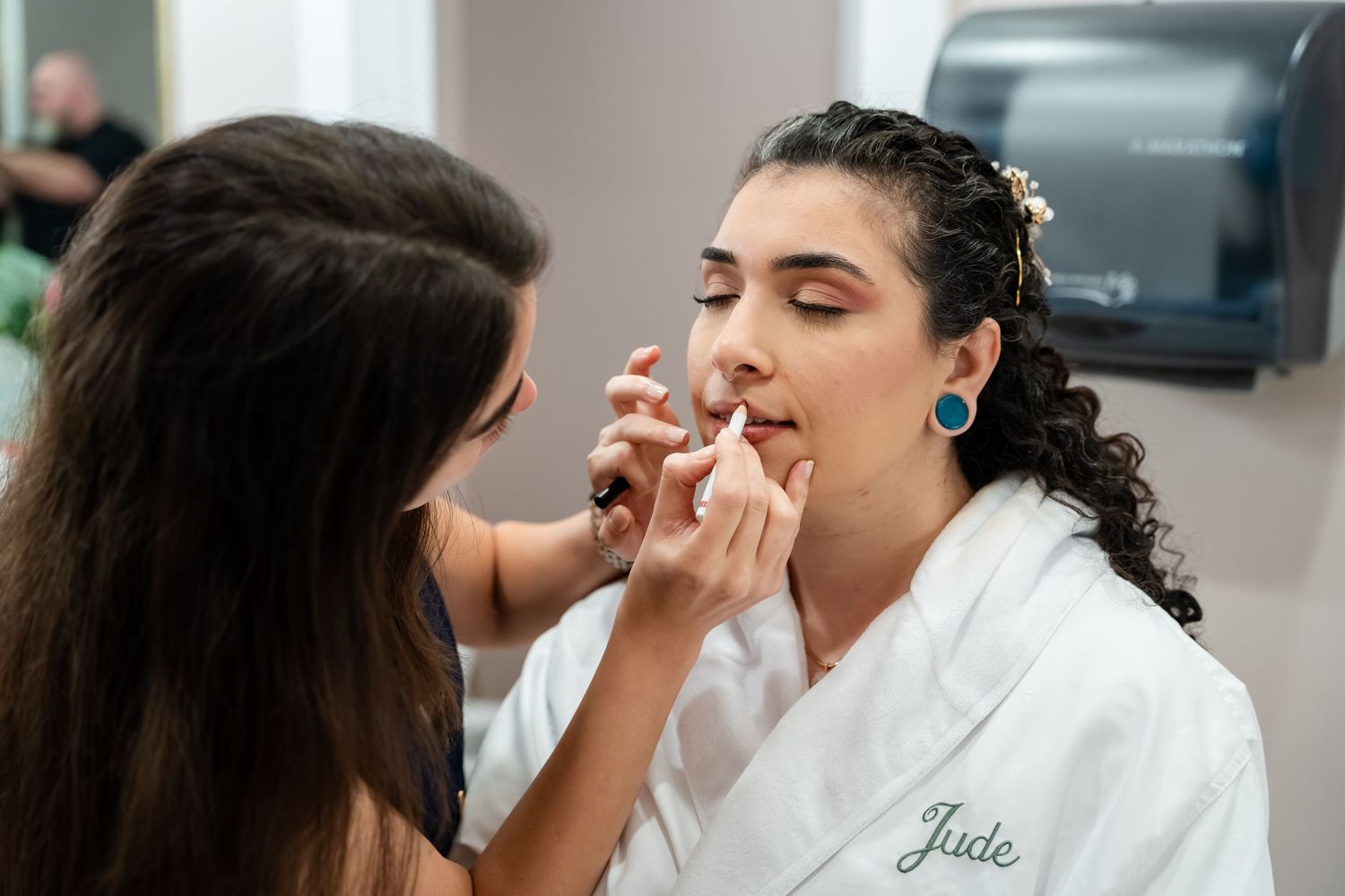 Makeup artist applying lipstick to a bride named Jade wearing a white robe during wedding preparations.
