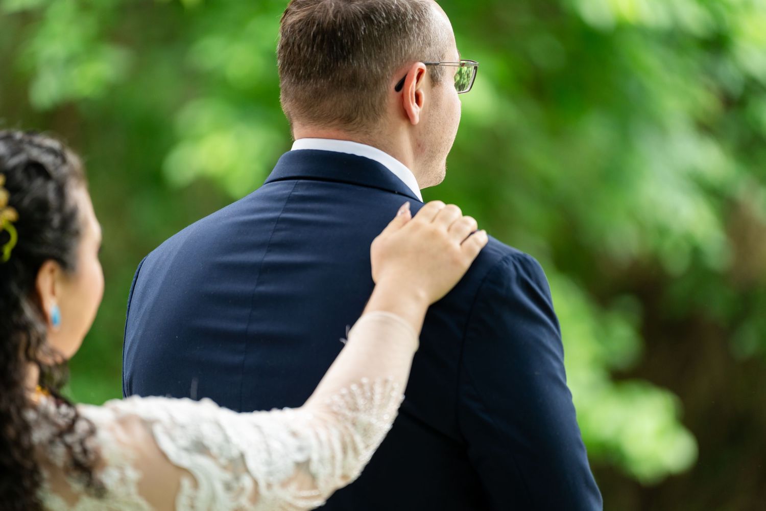 A bride in lace dress places hand on groom's shoulder in navy suit, standing outdoors among lush green trees.