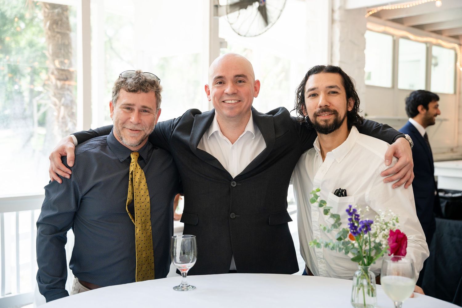 Three smiling men pose together at a bright restaurant event, one wearing a yellow tie, another in a white shirt.