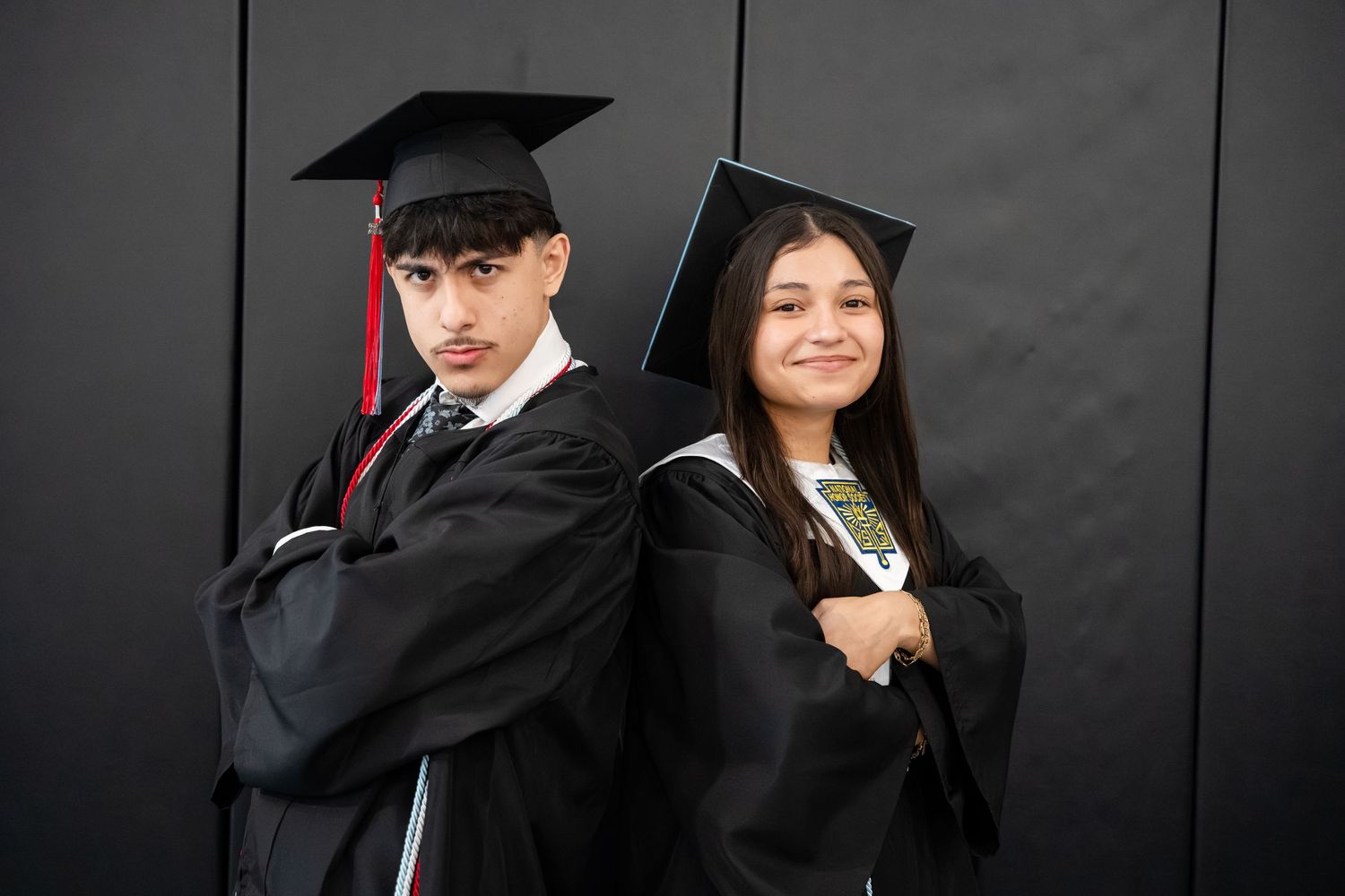 Two graduates in black caps and gowns pose back-to-back with arms crossed, smiling for a graduation photo.