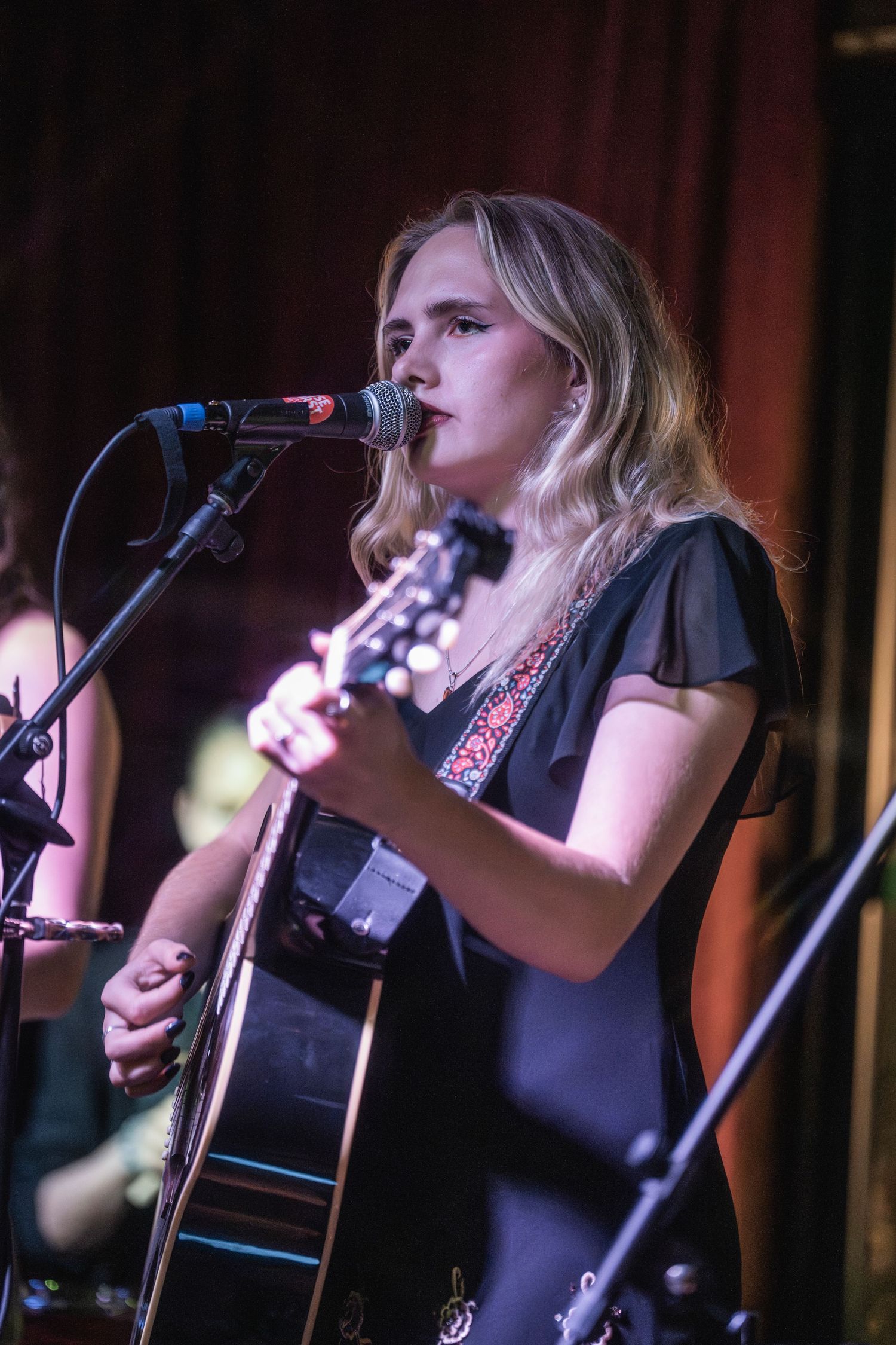 Female musician performs on acoustic guitar at dimly lit venue with microphone setup.
