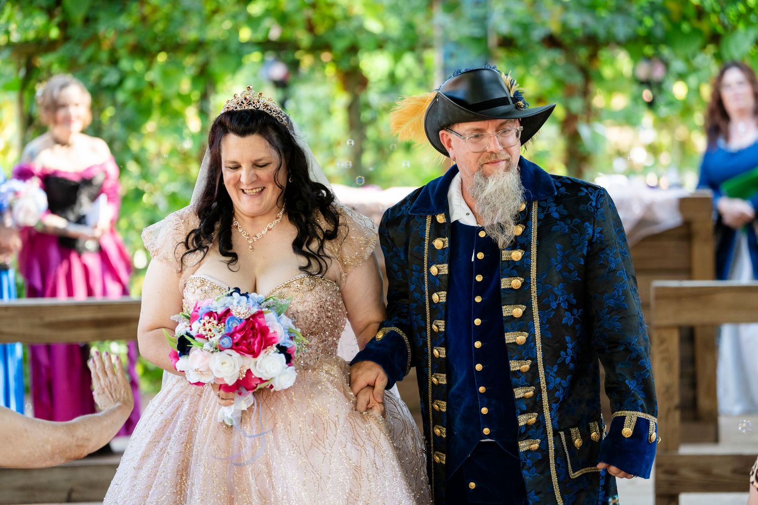 A couple walks down an outdoor wedding aisle surrounded by trees and guests in formal attire.