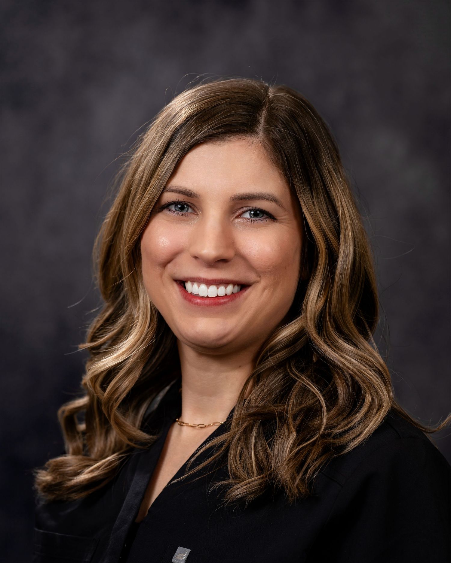 Professional headshot portrait of someone with wavy brown hair and highlights against a dark background.