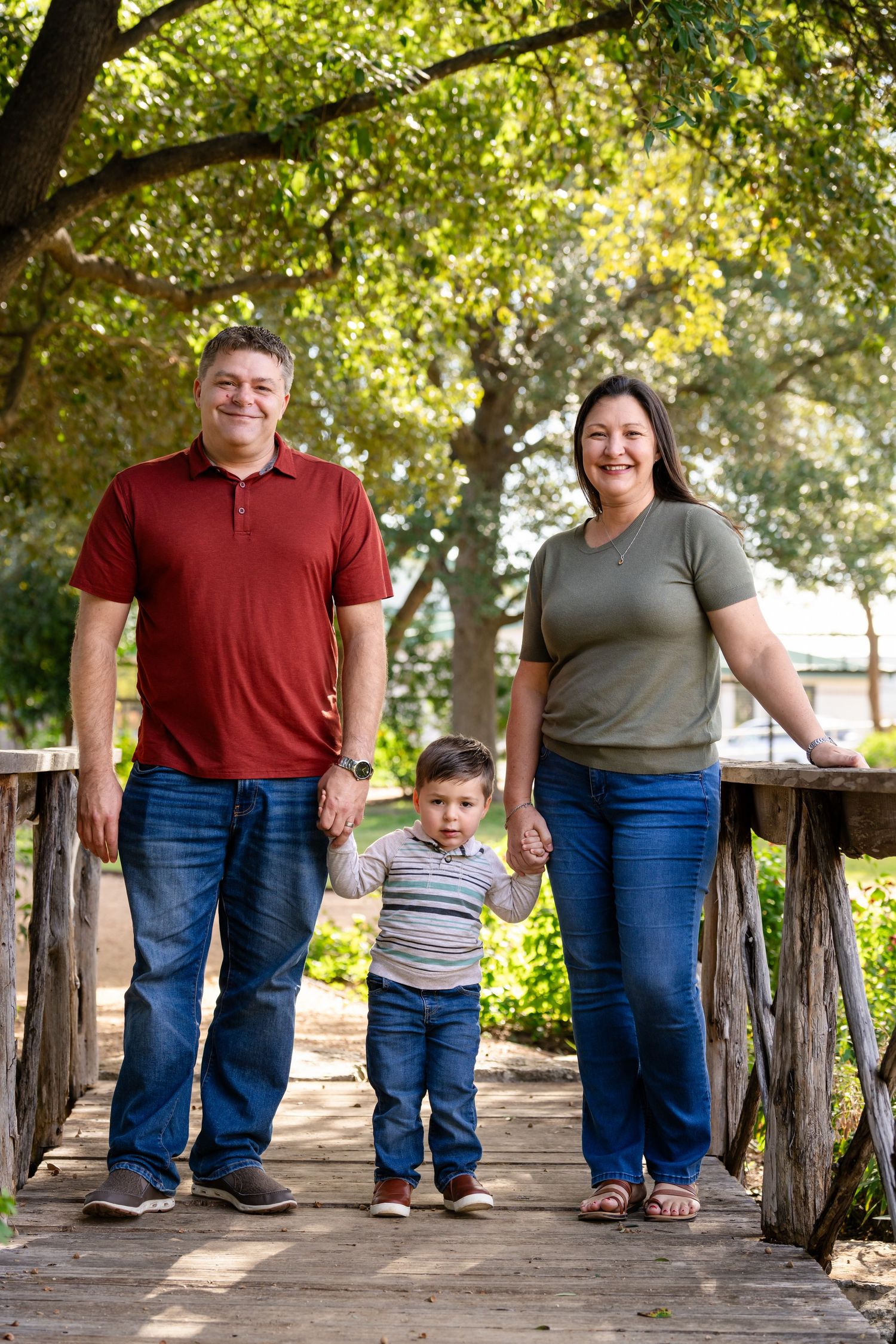 Family walking along rustic wooden fence path under sunlit green trees in outdoor park setting.