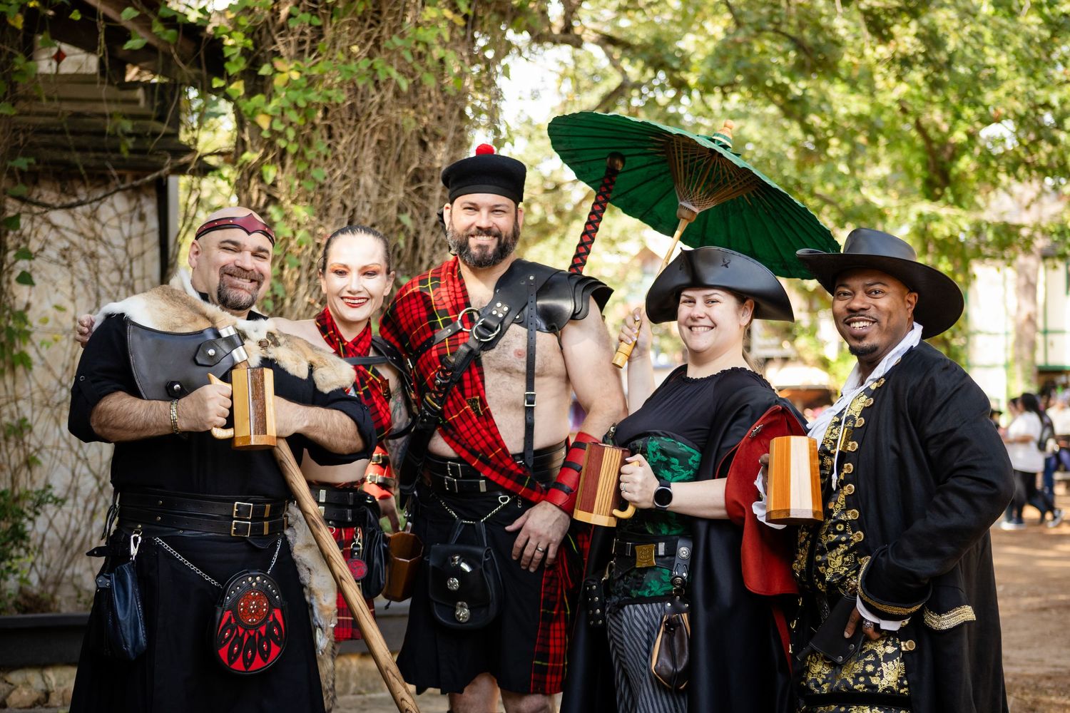 A group of performers in traditional Renaissance festival costumes with musical instruments pose together outdoors.