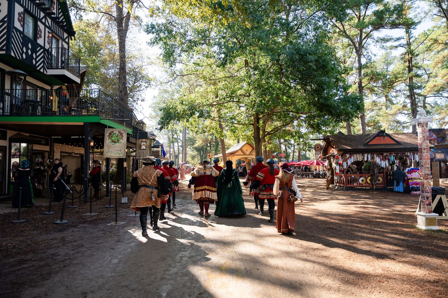 Panoramic view of a tree-lined outdoor marketplace with visitors browsing vendor stalls on a sunny day.