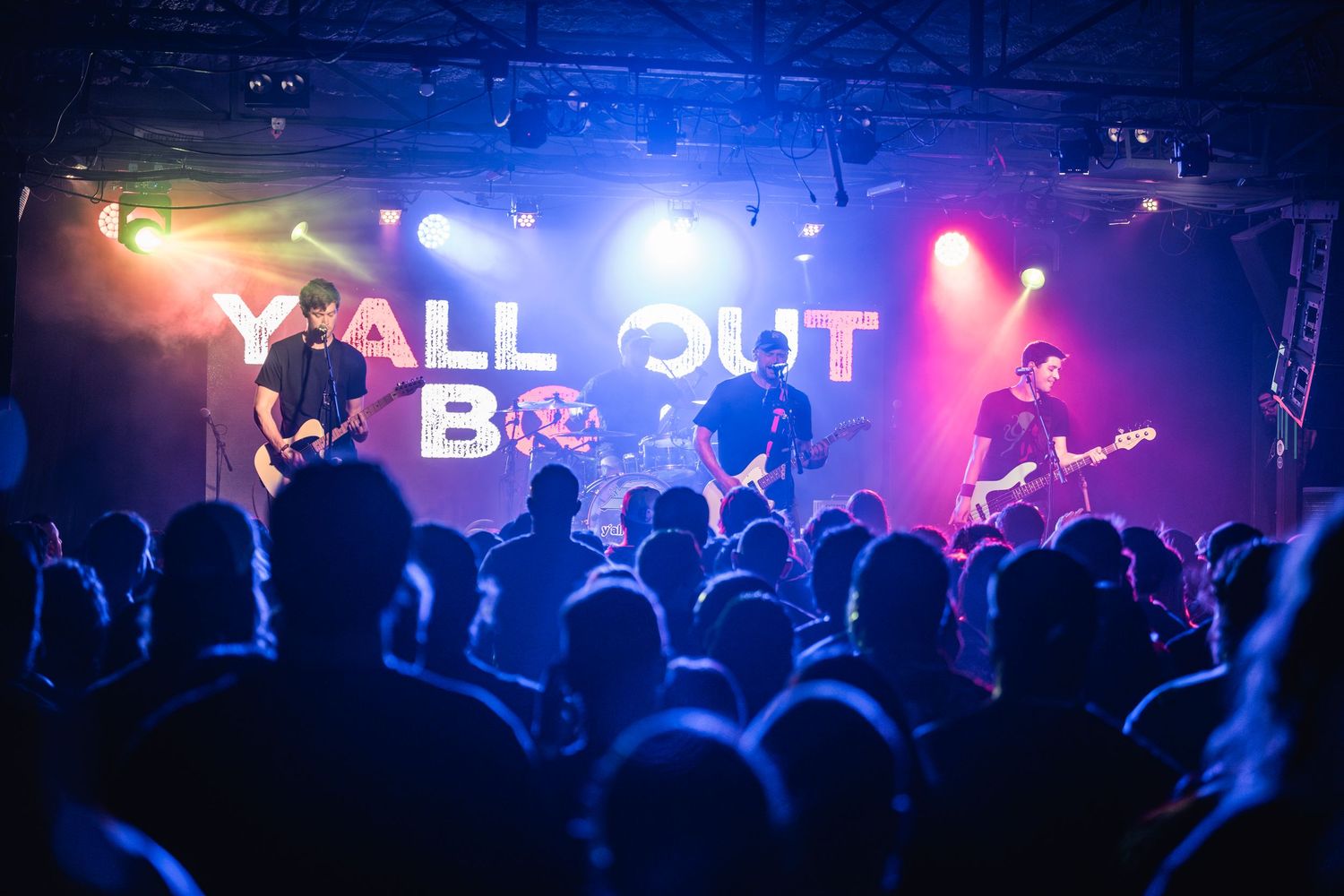 Concert crowd silhouettes with colorful stage lighting and neon text display during live music performance.