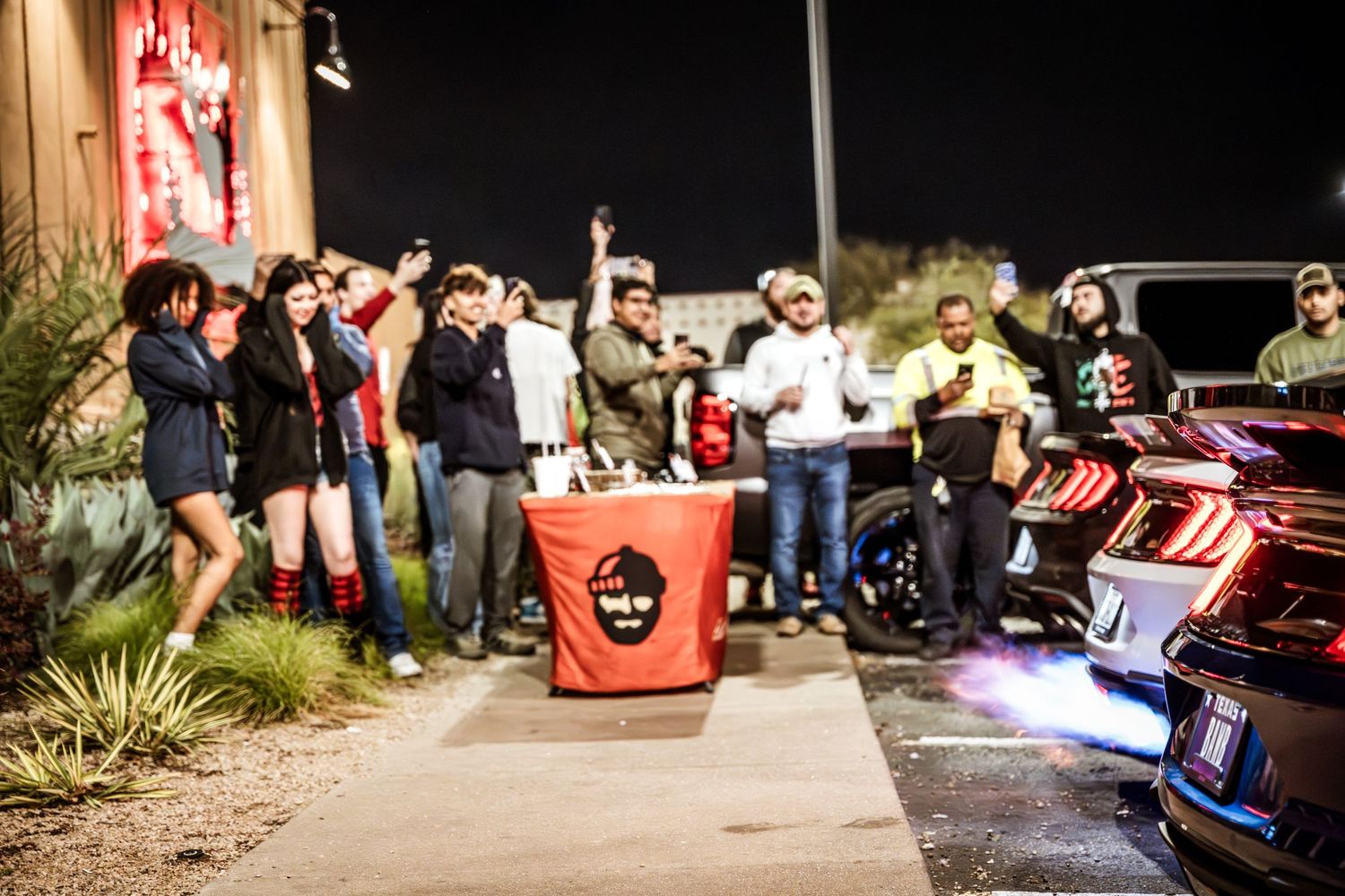 Large group of car enthusiasts gathered at night around modified vehicles with LED lighting.