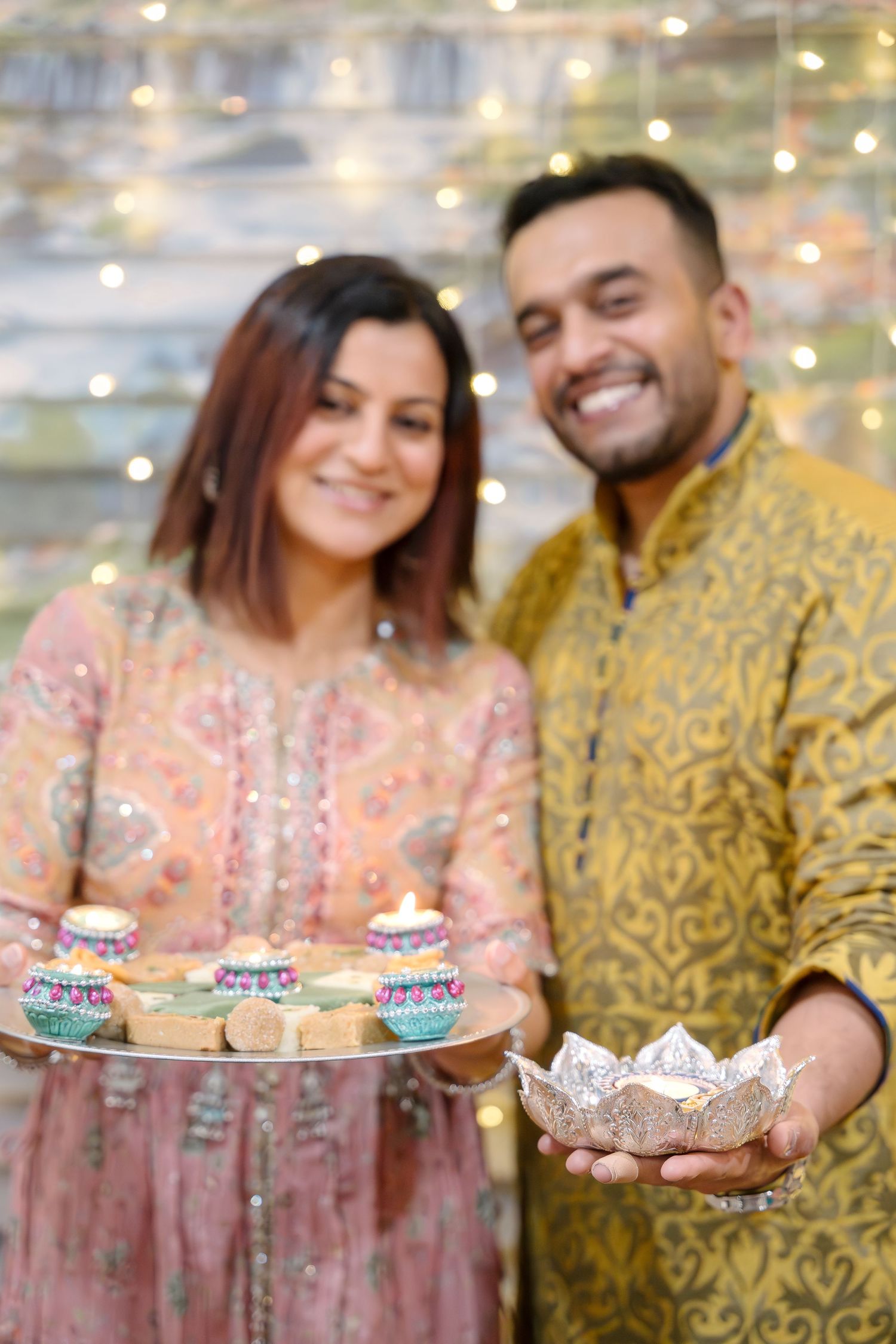 Couple in traditional Indian attire holding decorated sweets and treats at a celebration.