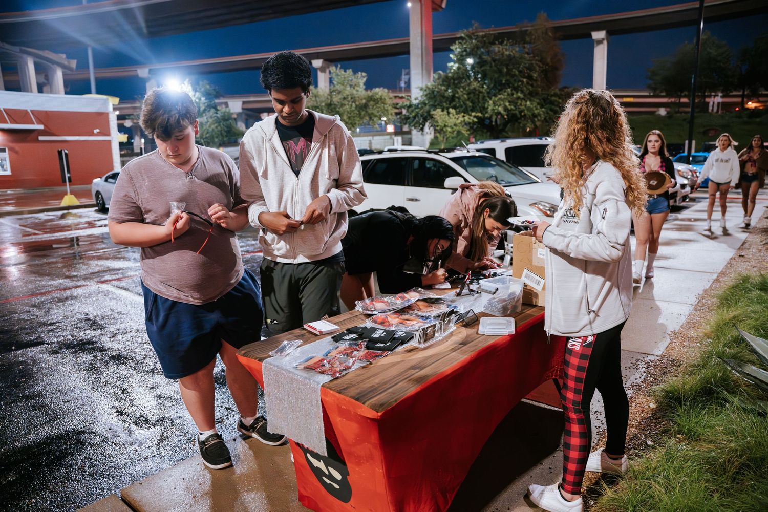 People browsing merchandise displayed on a table at an outdoor nighttime car meet.