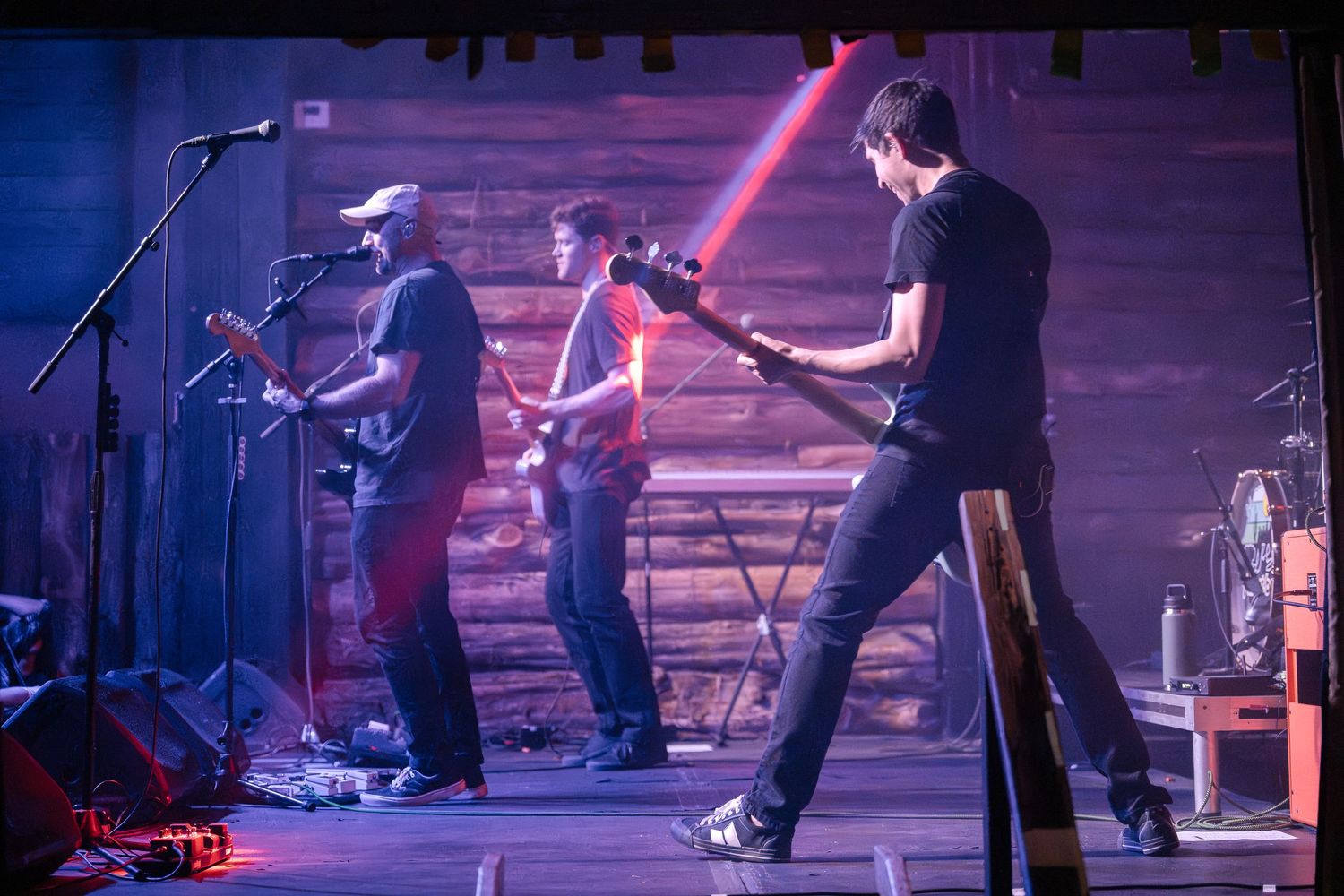 Musicians perform on stage with dramatic red lighting effects and wooden panel backdrop.