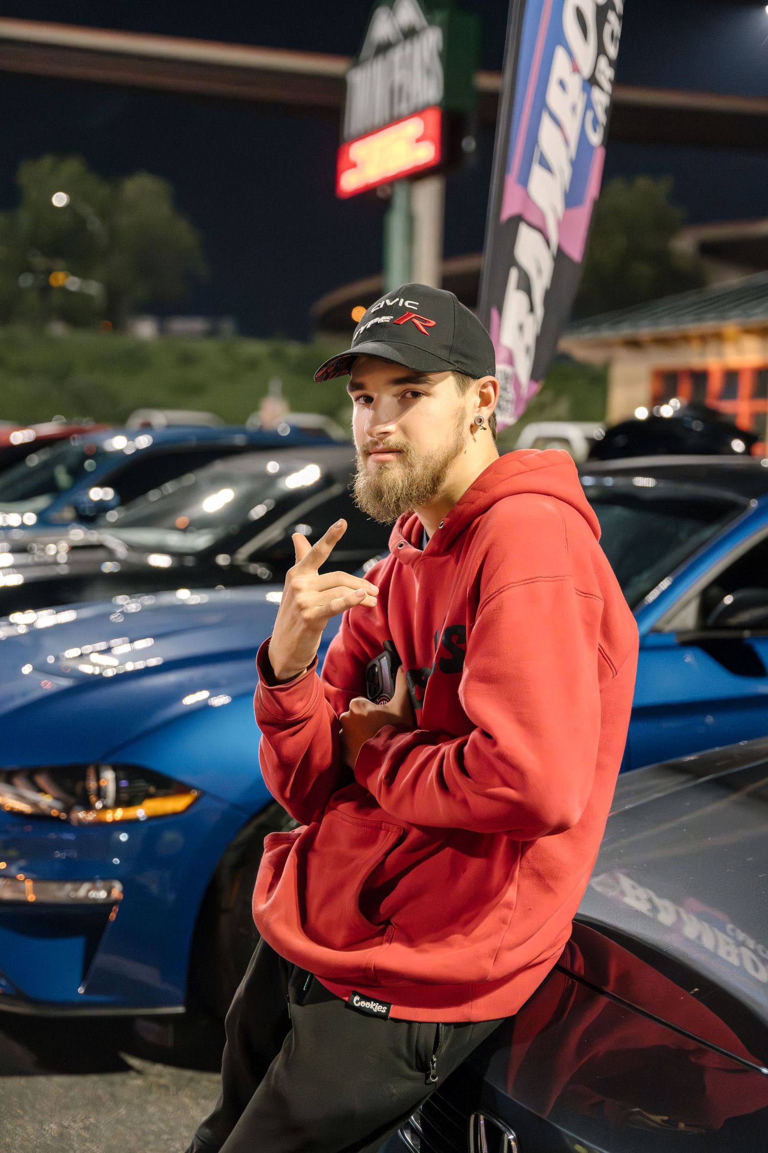 A casual car enthusiast in a red hoodie poses next to vehicles at a nighttime auto meetup.