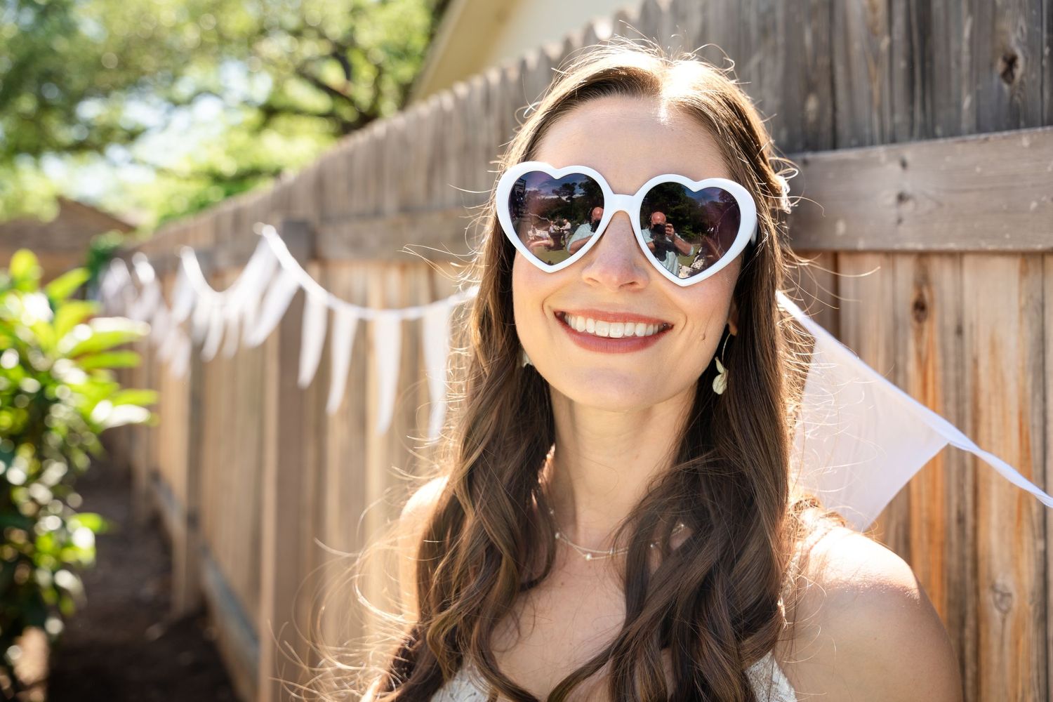 A close-up portrait wearing heart-shaped sunglasses with white bunting decoration in the background.