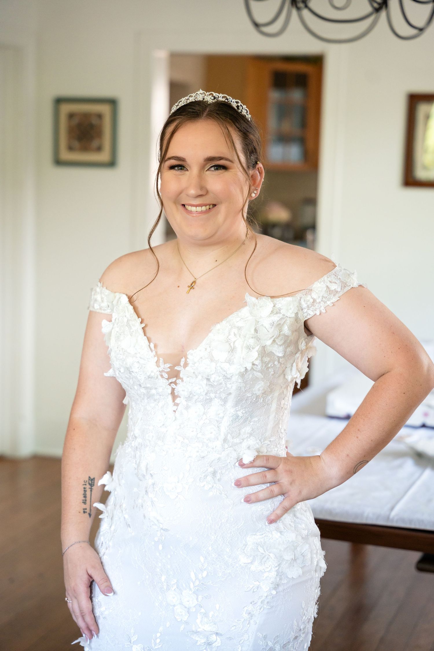 A bride poses in an elegant white lace wedding gown with off-shoulder design and delicate beading in an indoor setting.