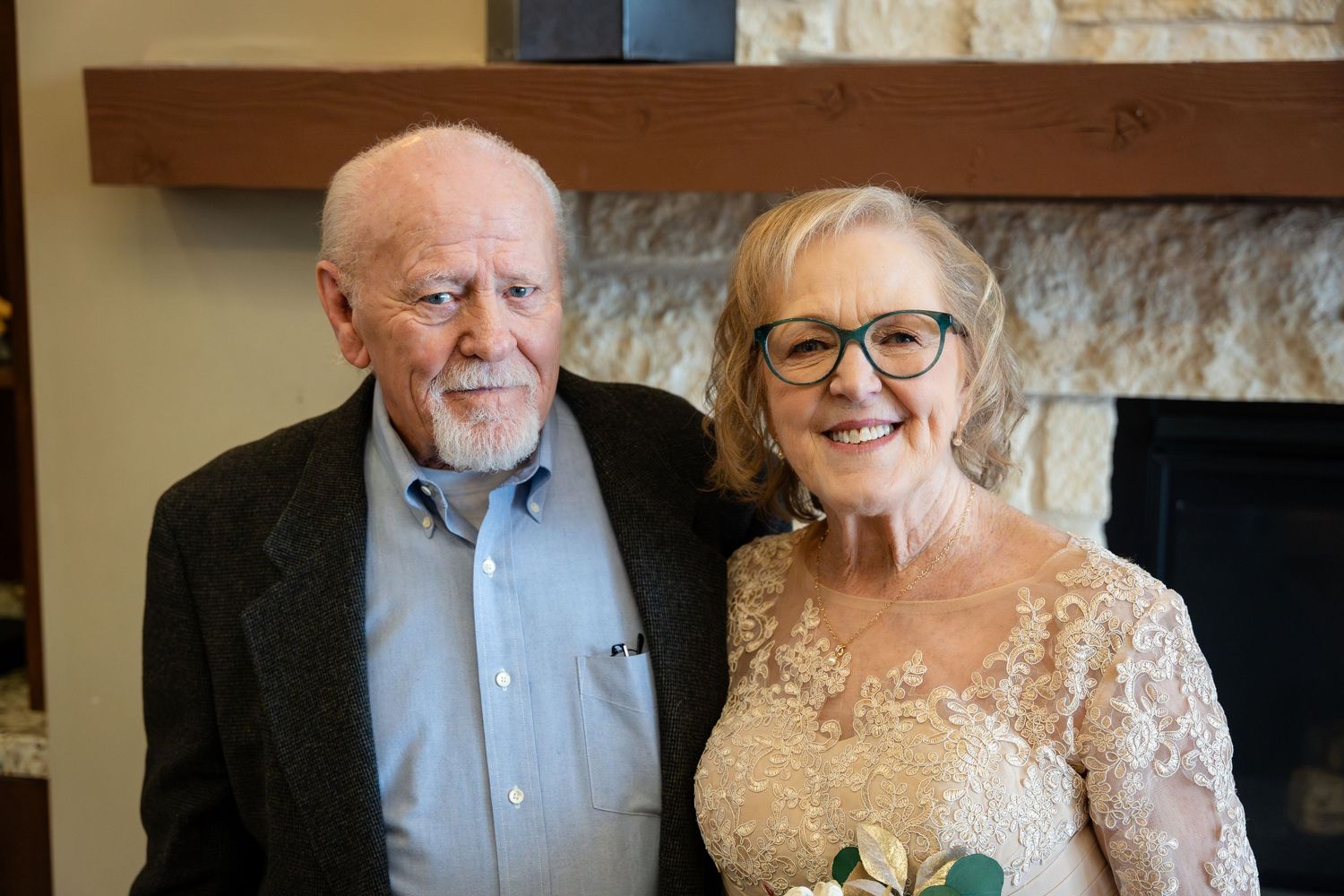 Two elegantly dressed elderly people in formal attire smile warmly together at an indoor venue by a fireplace.