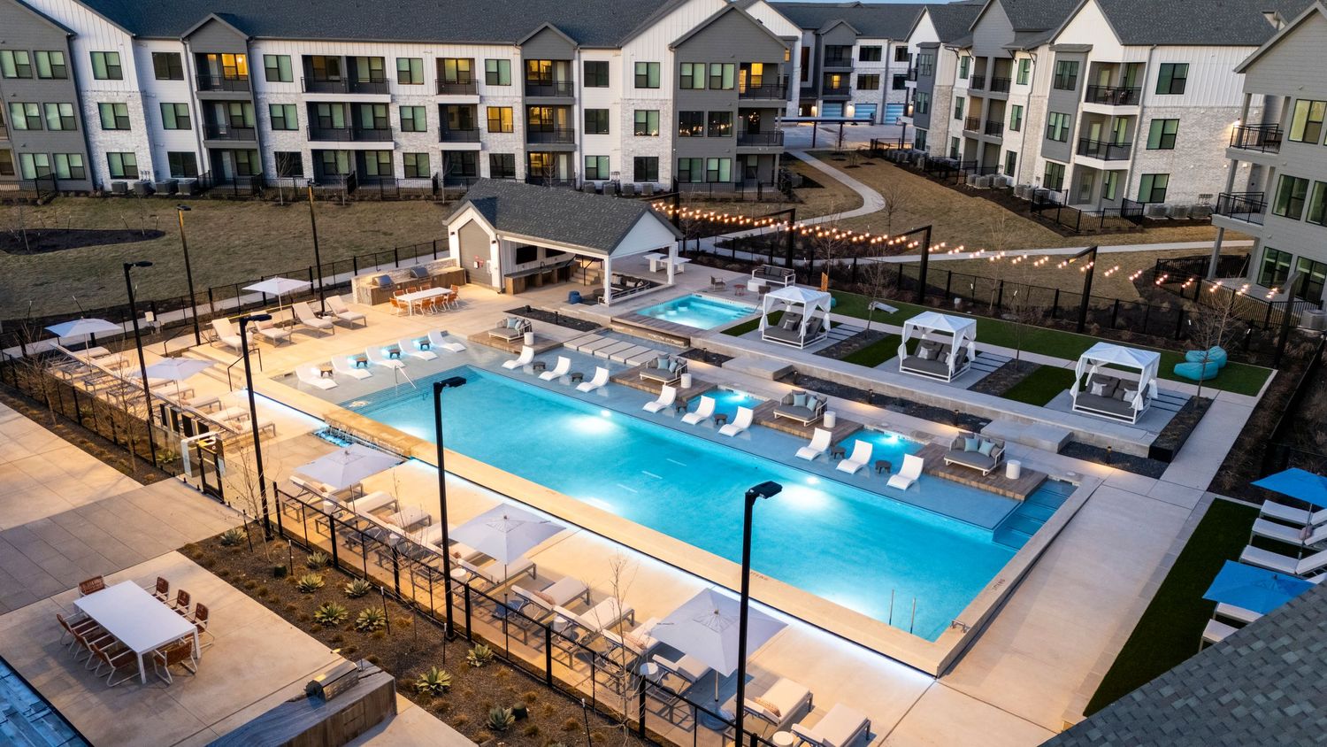 Illuminated luxury apartment complex swimming pool at dusk with lounge chairs and modern amenities.