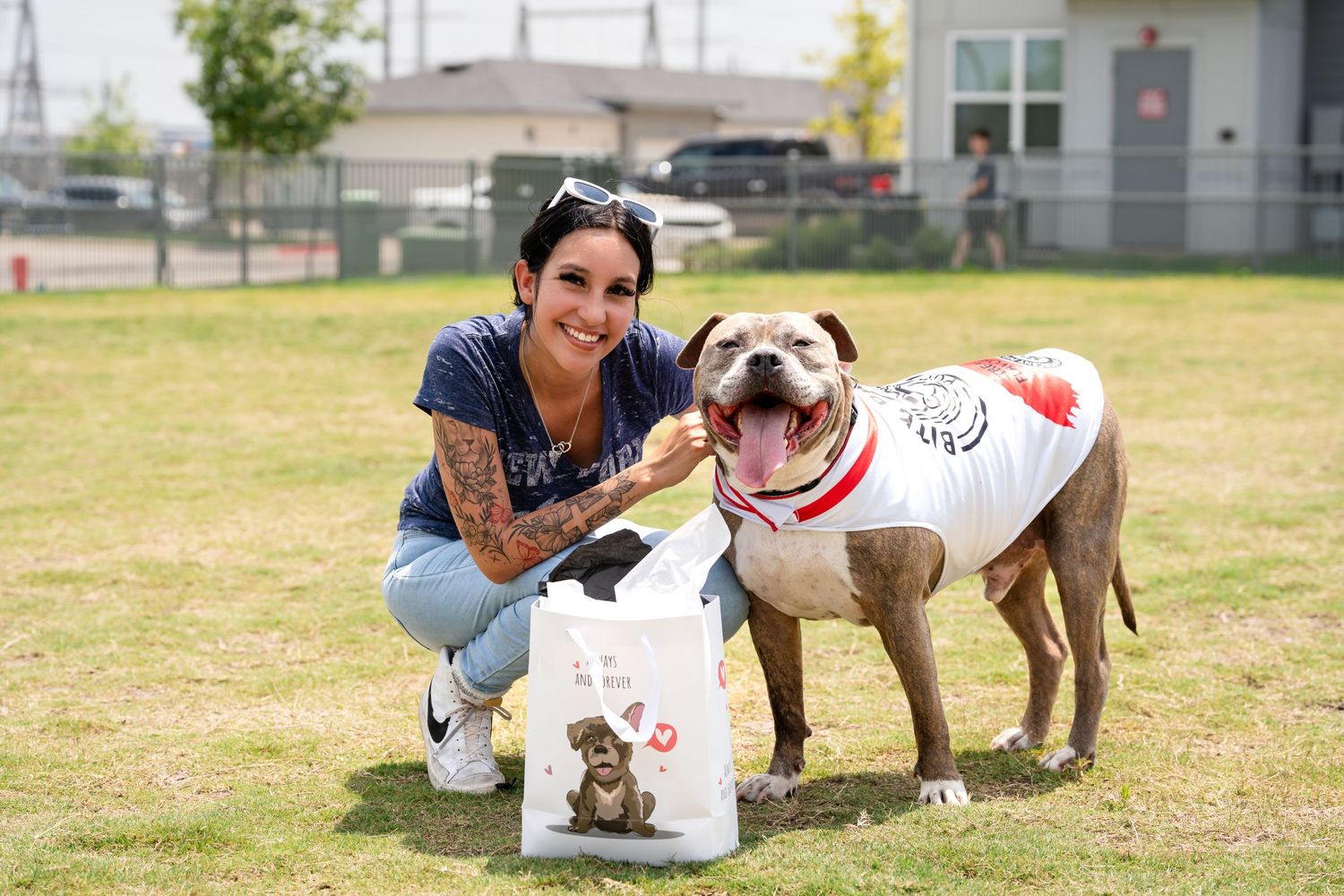 A happy pit bull wearing a white shirt poses with their owner and pet supplies on grass.