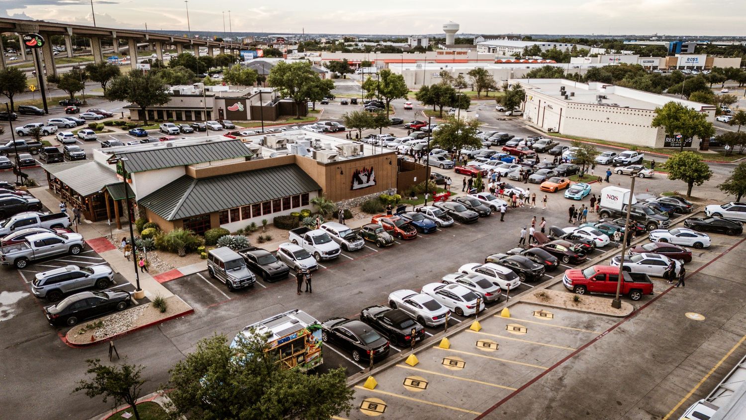 Aerial view of crowded parking lot with retail buildings and parked vehicles.