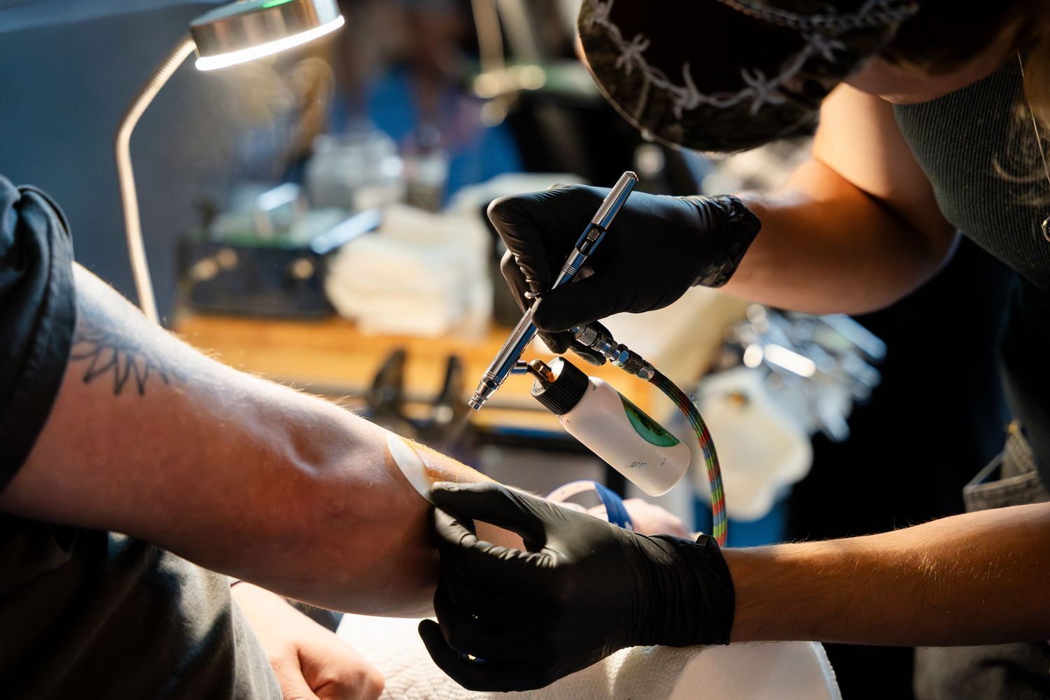 A tattoo artist wearing black gloves works on a client's arm with a professional tattoo machine.