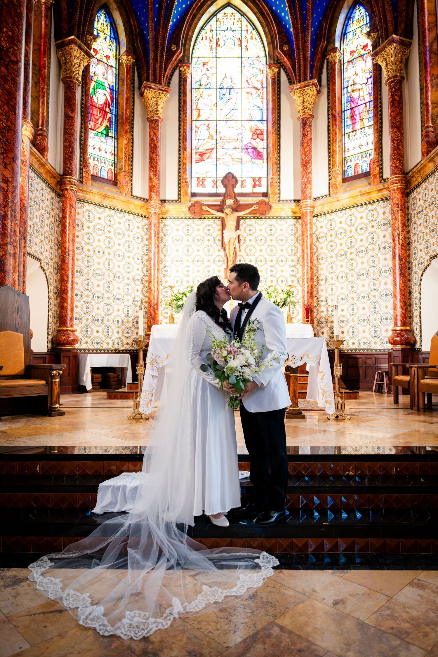 Newlyweds share a romantic kiss in a stunning cathedral with stained glass windows and ornate altar decor.