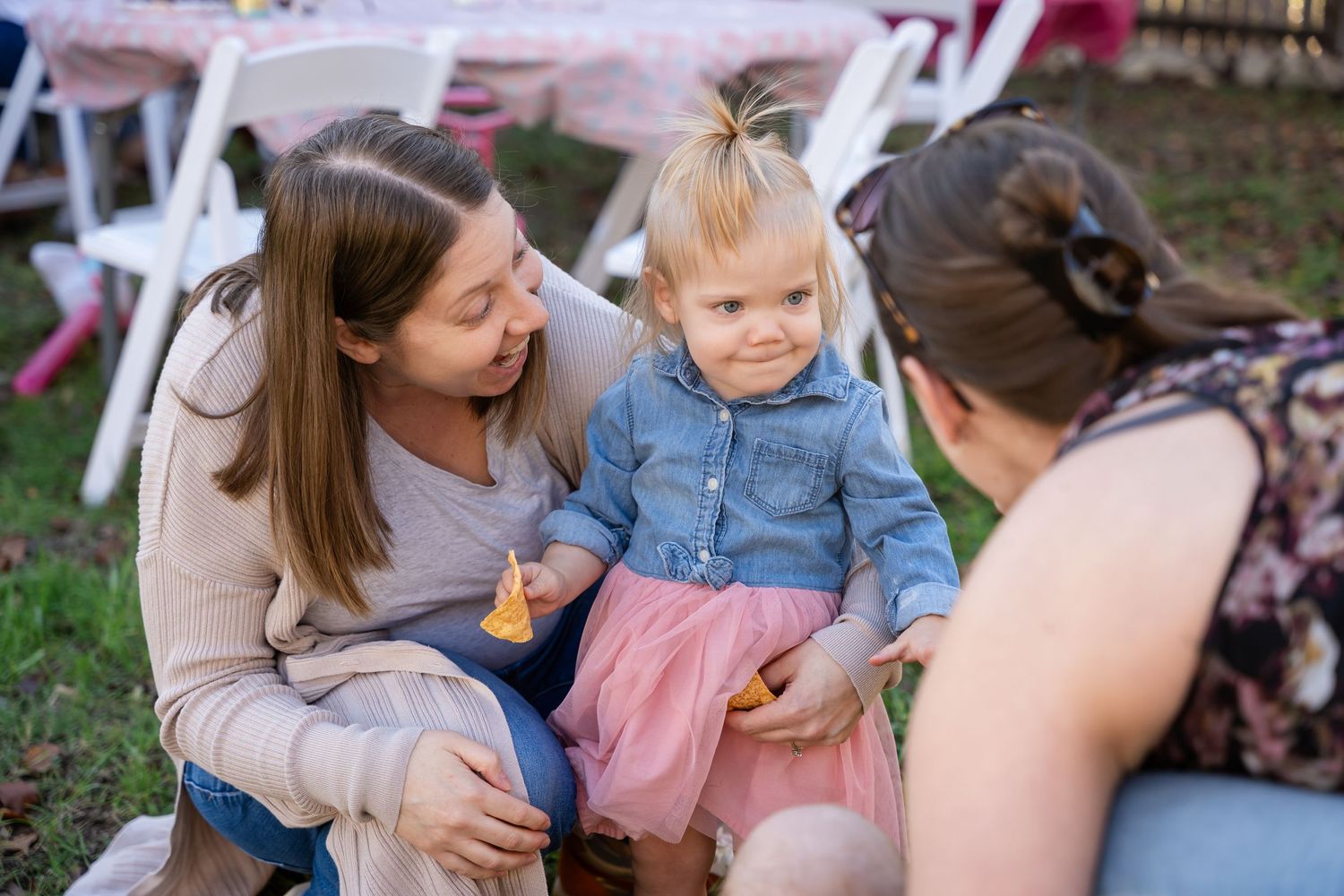 A small child in a denim shirt and pink tulle skirt sits outdoors between two adults on a sunny day.