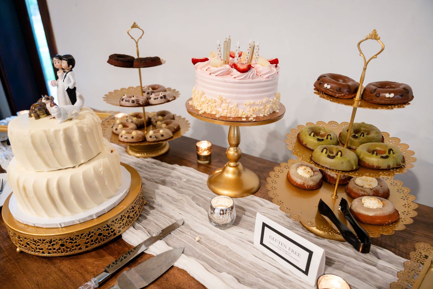 White wedding cake with simple design on gold cake stand, surrounded by cupcakes and cookies on a dessert table display.