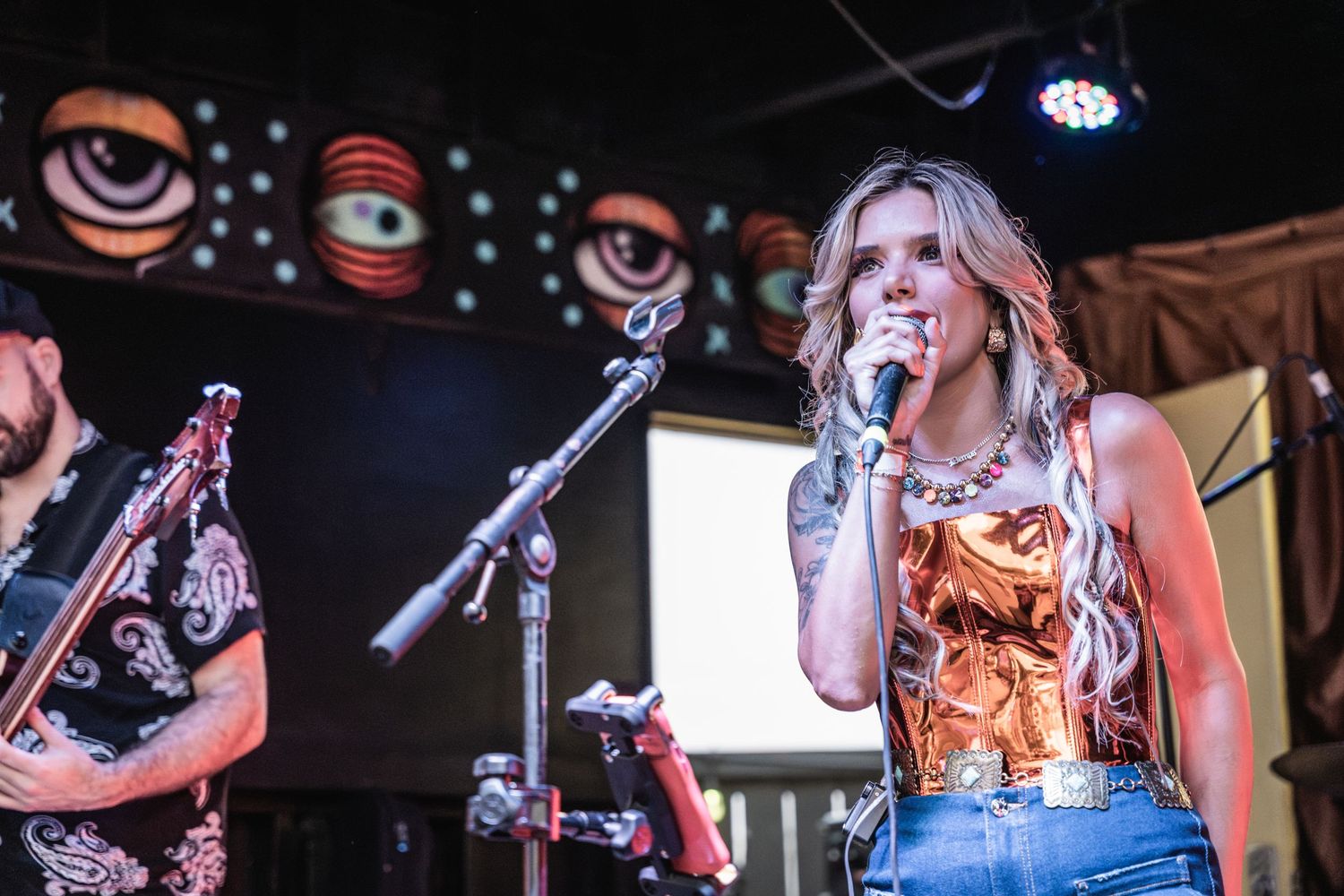 Singer performs on stage in orange corset top and jeans with band members and eye-themed backdrop decor.